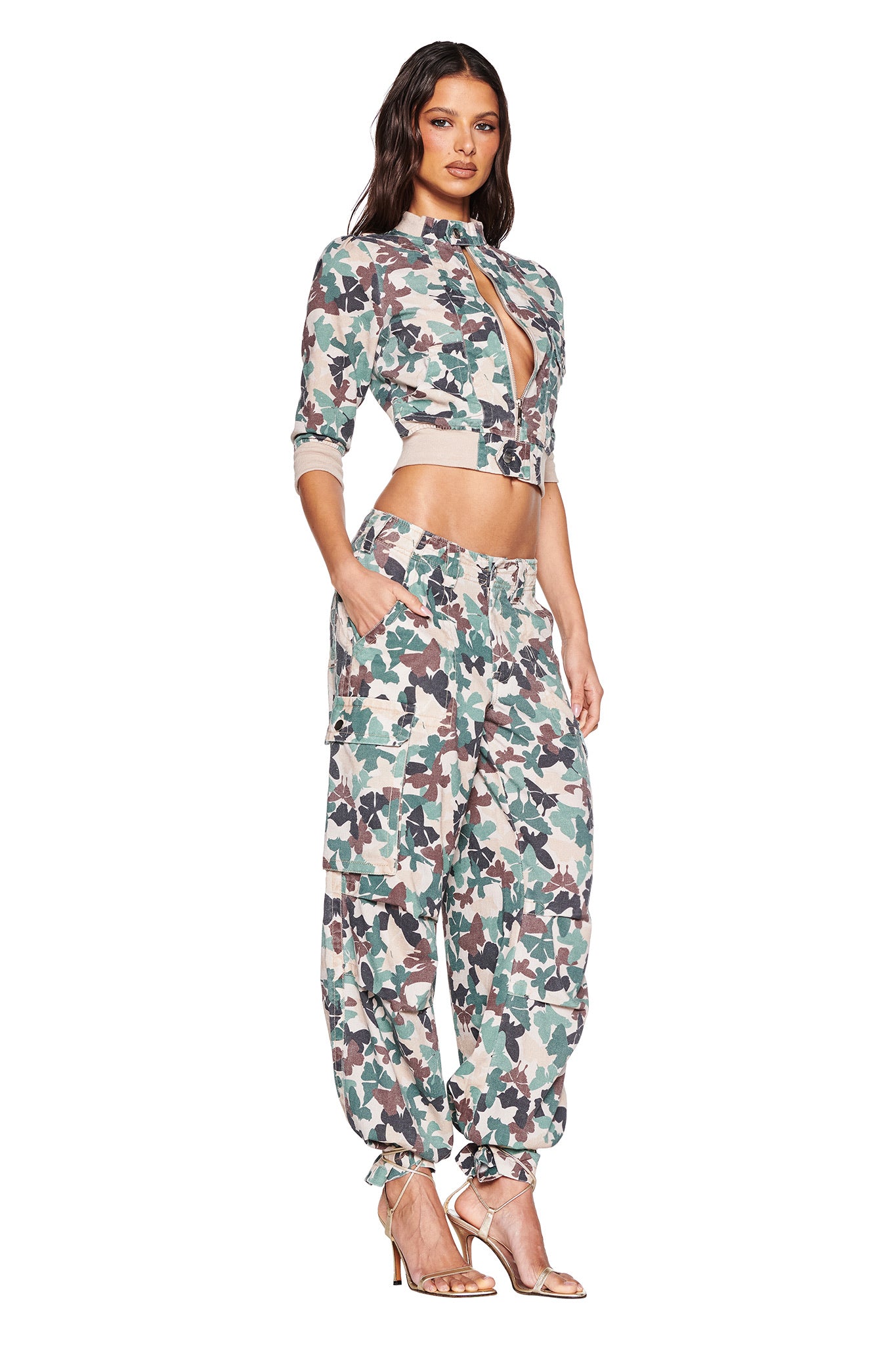 A woman poses elegantly in I.AM.GIA's EFFIE CARGO PANT - FLUTTER CAMO with a cropped jacket. Crafted from lightweight linen, the high-waisted pants blend green, brown, and beige hues on a white background as she stands sideways with hands in pockets and perched on high heels.