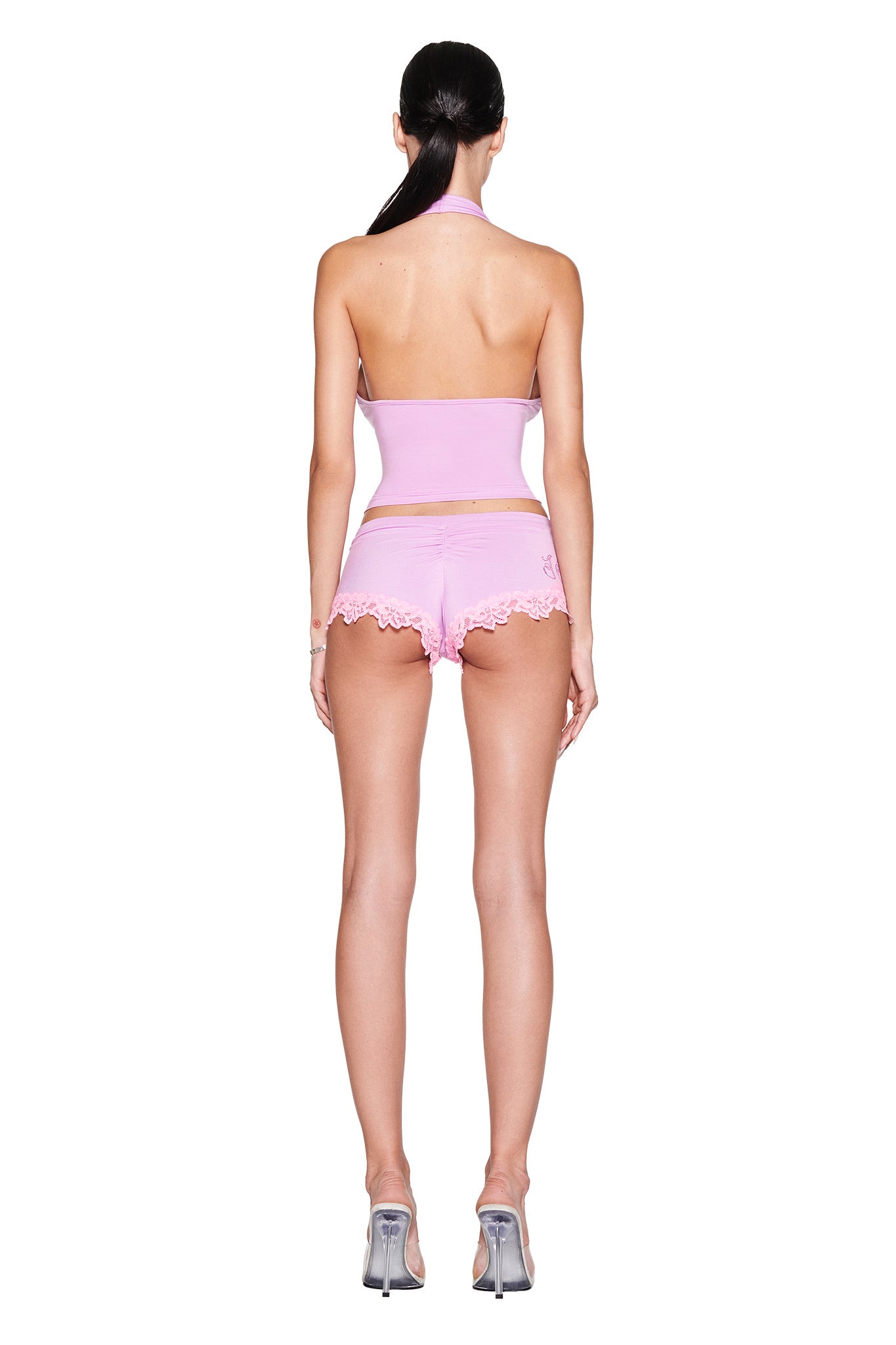 A person with long dark hair wears the lavender CELESTE SHORT and a matching pink halter top from I.AM.GIA, showcasing a bodycon silhouette. They stand on a reflective surface in silver high heels against a plain white background.