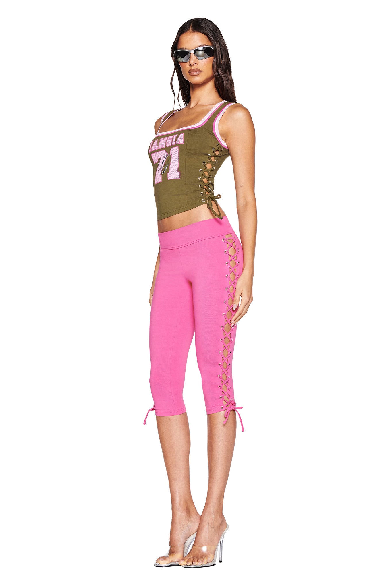 A woman in silver heels poses against a white backdrop in sunglasses, wearing the I.AM.GIA NOA TOP - GREEN with "Mangia 71" and pink capri pants featuring lace-up side details, adding flair to her outfit.