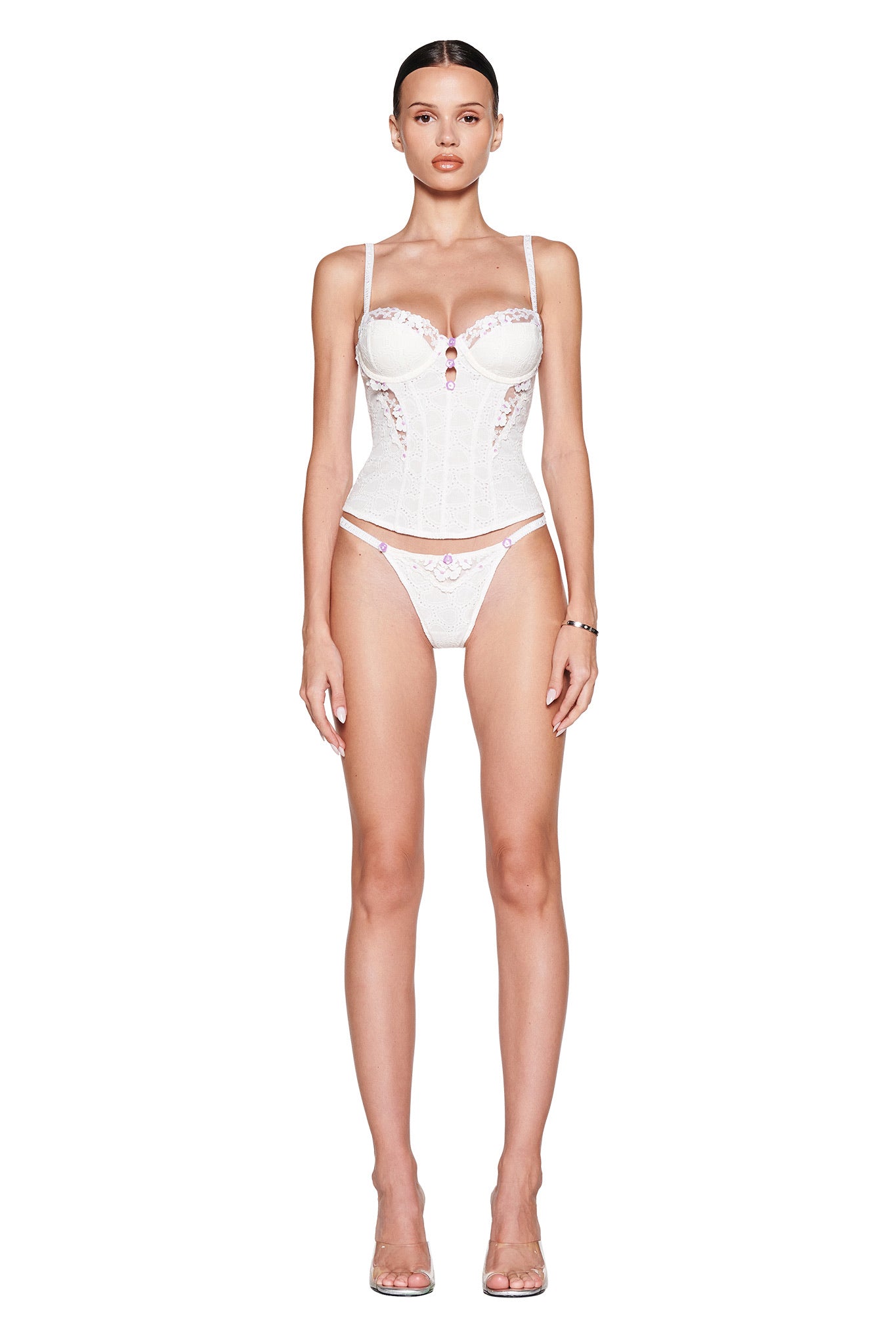 A woman faces forward in the I.AM.GIA ELLA BUSTIER SET - WHITE, featuring a white lace cotton bustier with sheer embroidered panels and matching underwear. She wears clear heels and has her dark hair pulled back against a plain white background.