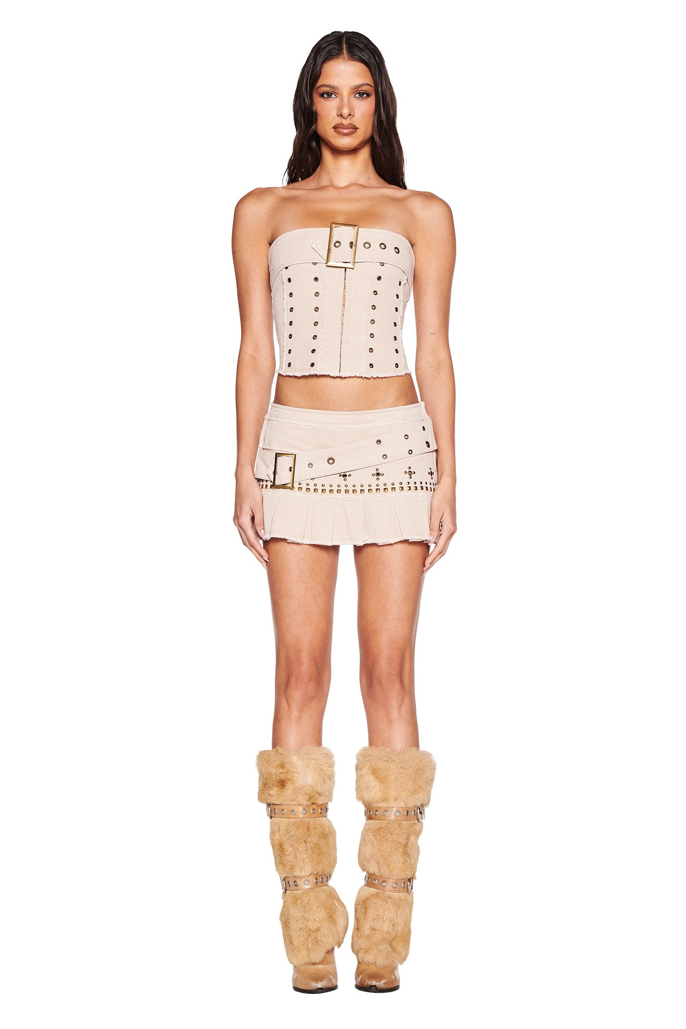 A woman faces forward in the I.AM.GIA MASON MINI SKIRT - TAN with metallic buckle and grommet details, styled with a matching strapless top, asymmetrical belt, tan faux-fur leg warmers, and high heels against a white background.