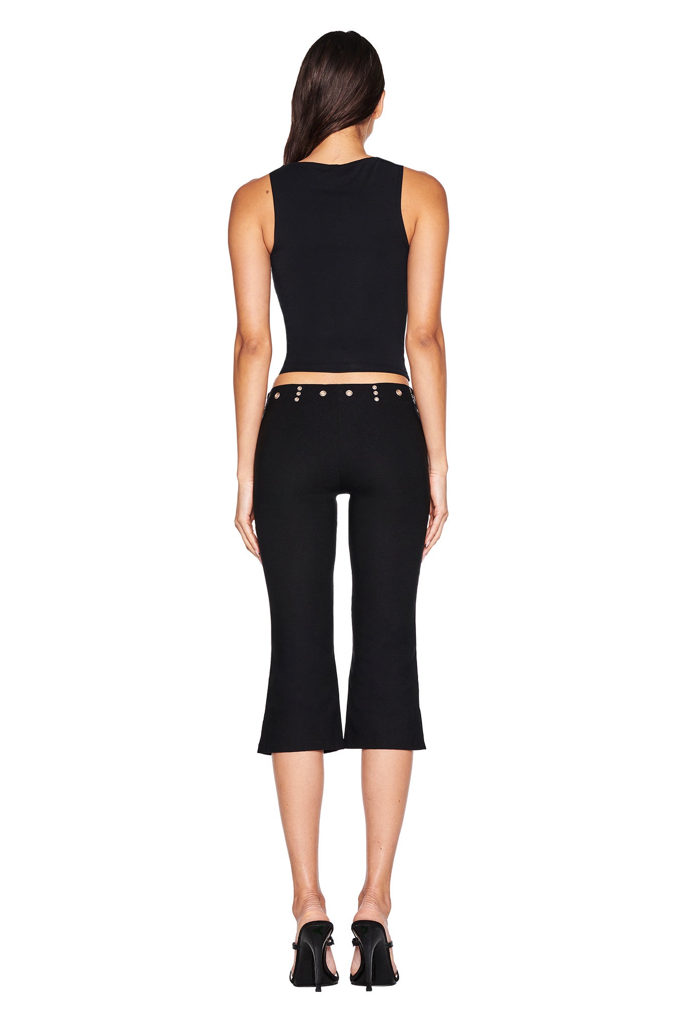 A woman faces away in a black sleeveless top, SHARAH CAPRI - BLACK slim-fit capri pants with silver grommets by I.AM.GIA, and black high heels. White background.