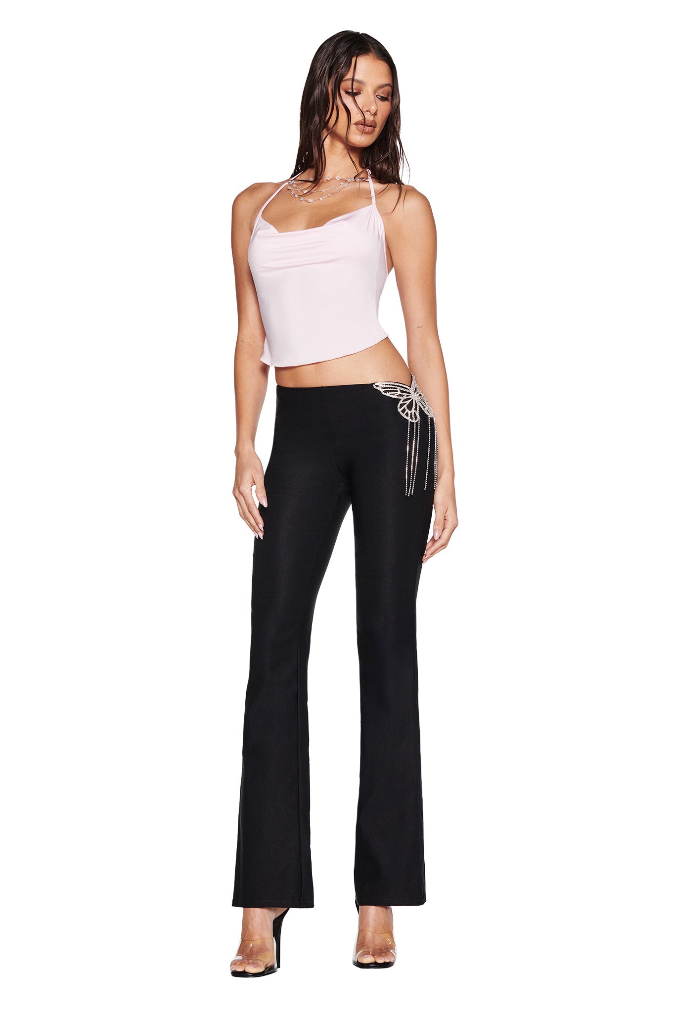 A woman stands against a white background wearing the I.AM.GIA SORREL PANT in black, featuring mid rise flared legs and dangling crystal chains at the hip, styled with a light pink camisole and clear high-heeled shoes.
