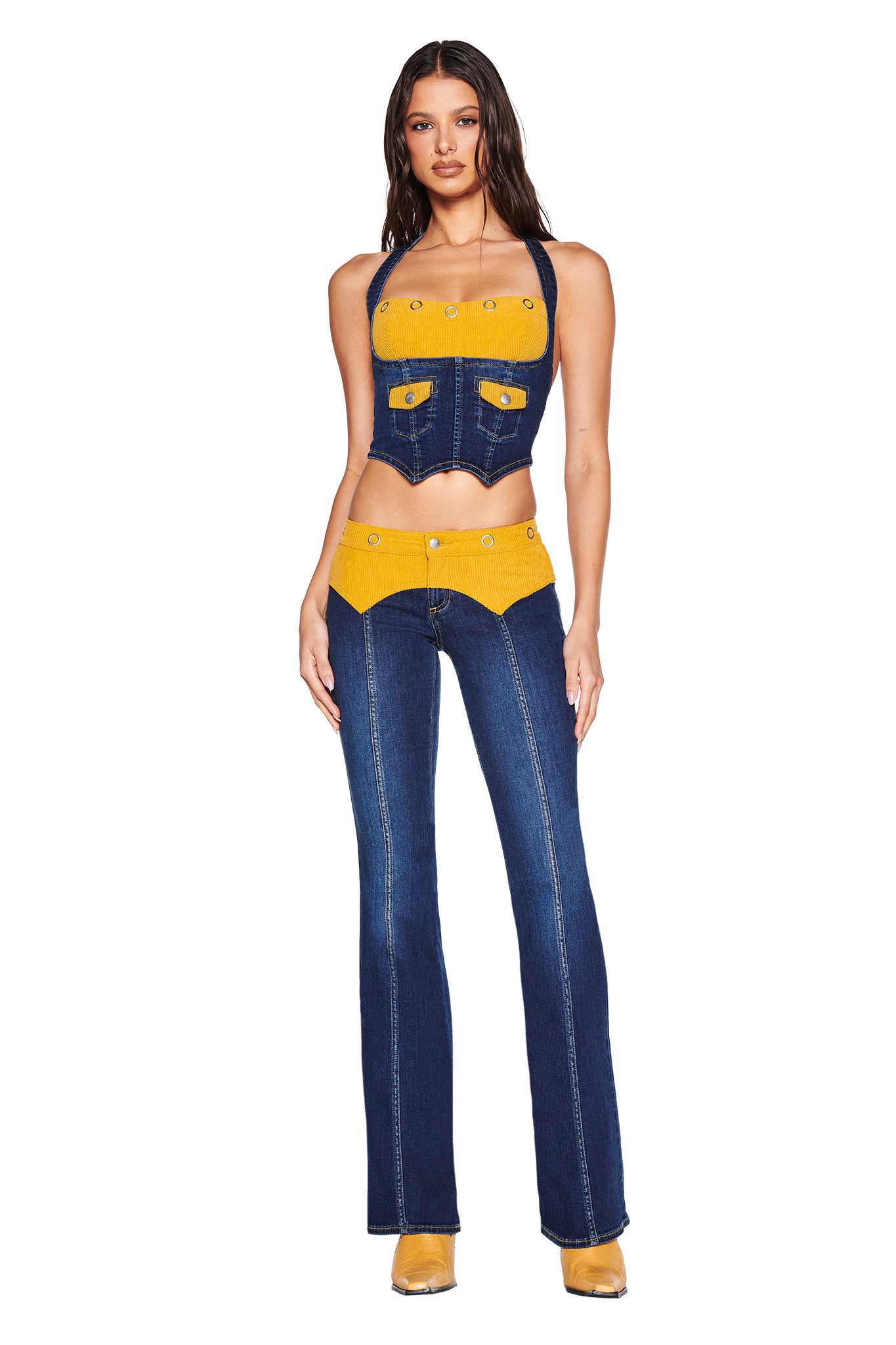 A woman faces forward in the I.AM.GIA TOMMIE JEAN - DARK WASH set, wearing a mid-rise denim halter top and flared pants with yellow accents, bold stitching, button details, and GIA embroidery. She has long wavy hair and yellow shoes.