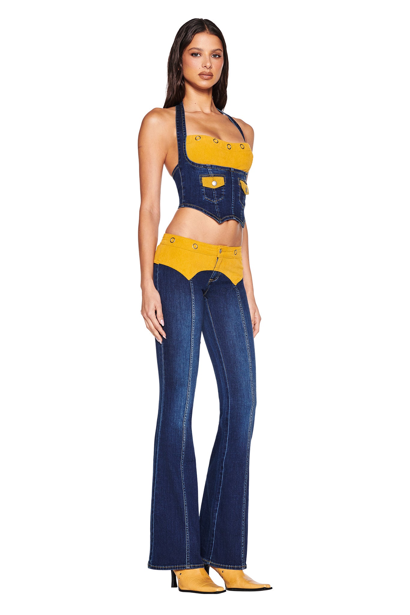 A woman models the I.AM.GIA TOMMIE JEAN - DARK WASH set: a cropped halter denim top and matching flared pants with a mustard yellow corduroy waistband and GIA logo, styled with yellow heeled boots against a white background.