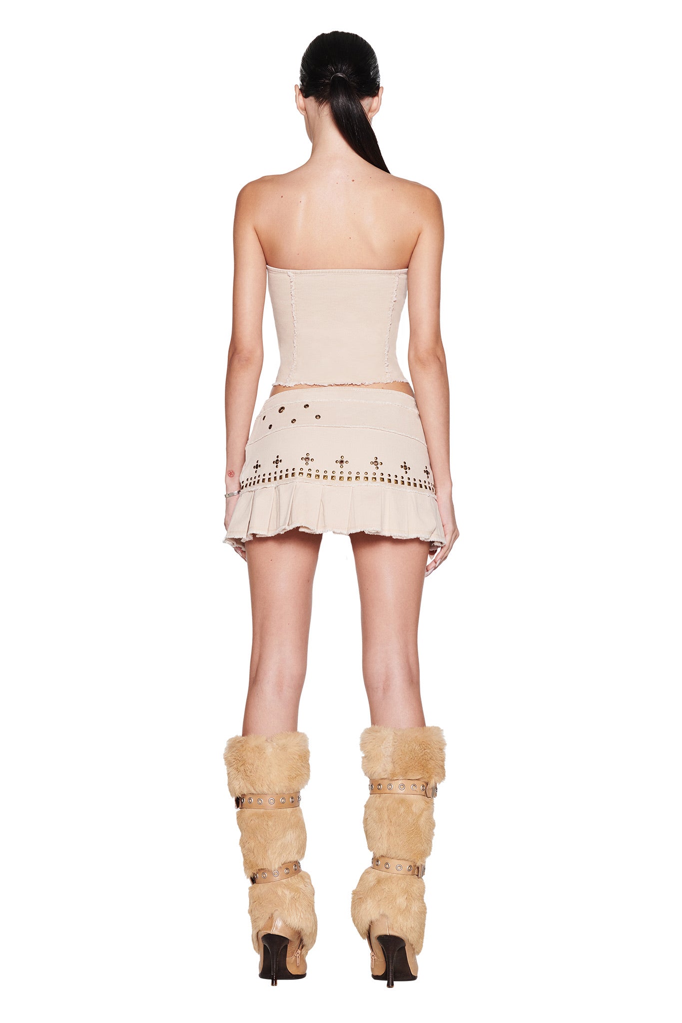 A woman with long dark hair in a ponytail stands facing away, wearing the I.AM.GIA MASON CORSET - TAN mini dress with strapless cutout details and pleats, paired with tall tan fur boots featuring straps and heels.