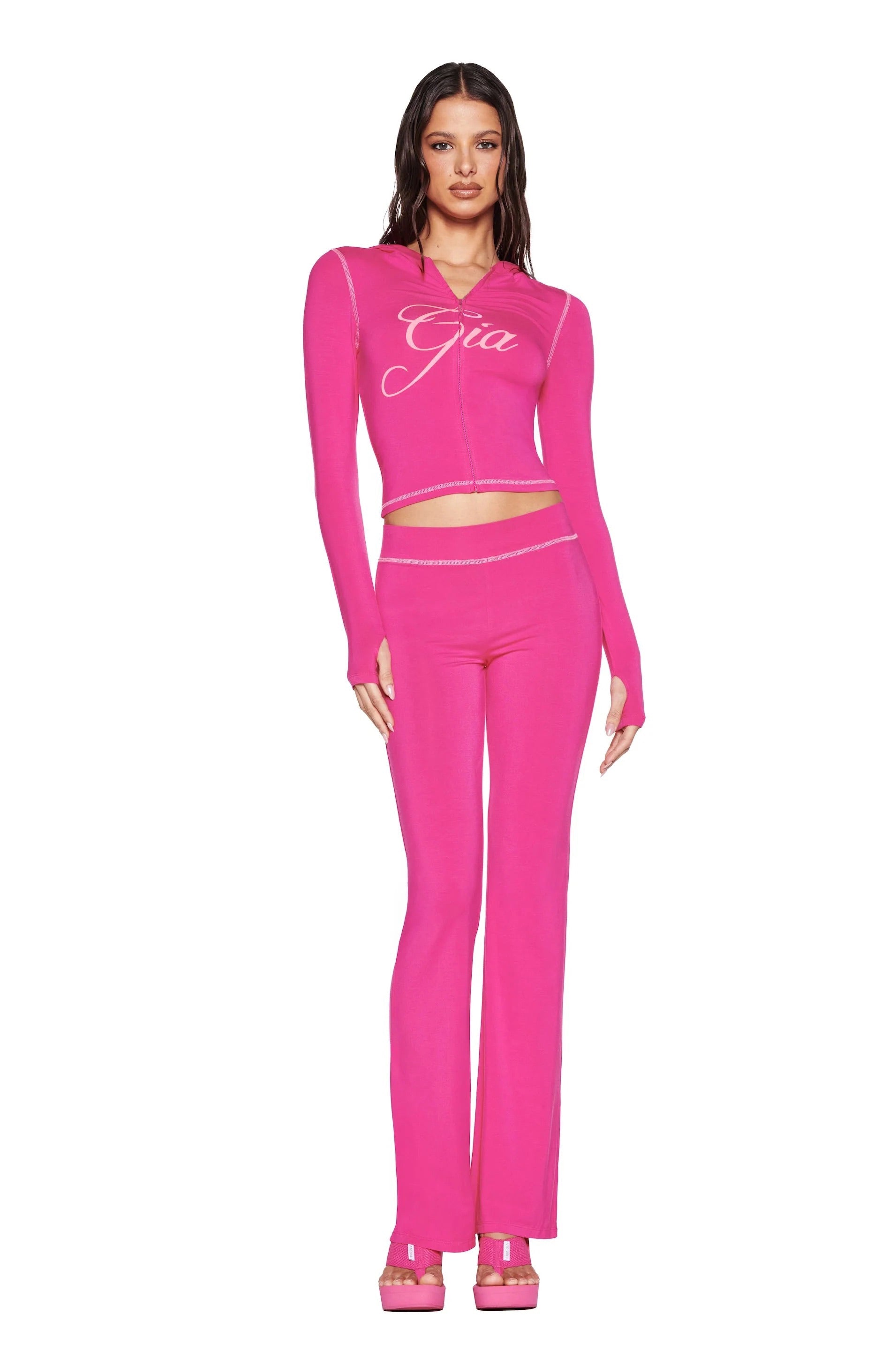 A woman poses against a white background wearing I.AM.GIA’s BLARE TRACKPANT in hot pink, styled with flared pants, a long-sleeve crop top featuring "Gia," and matching platform heels.