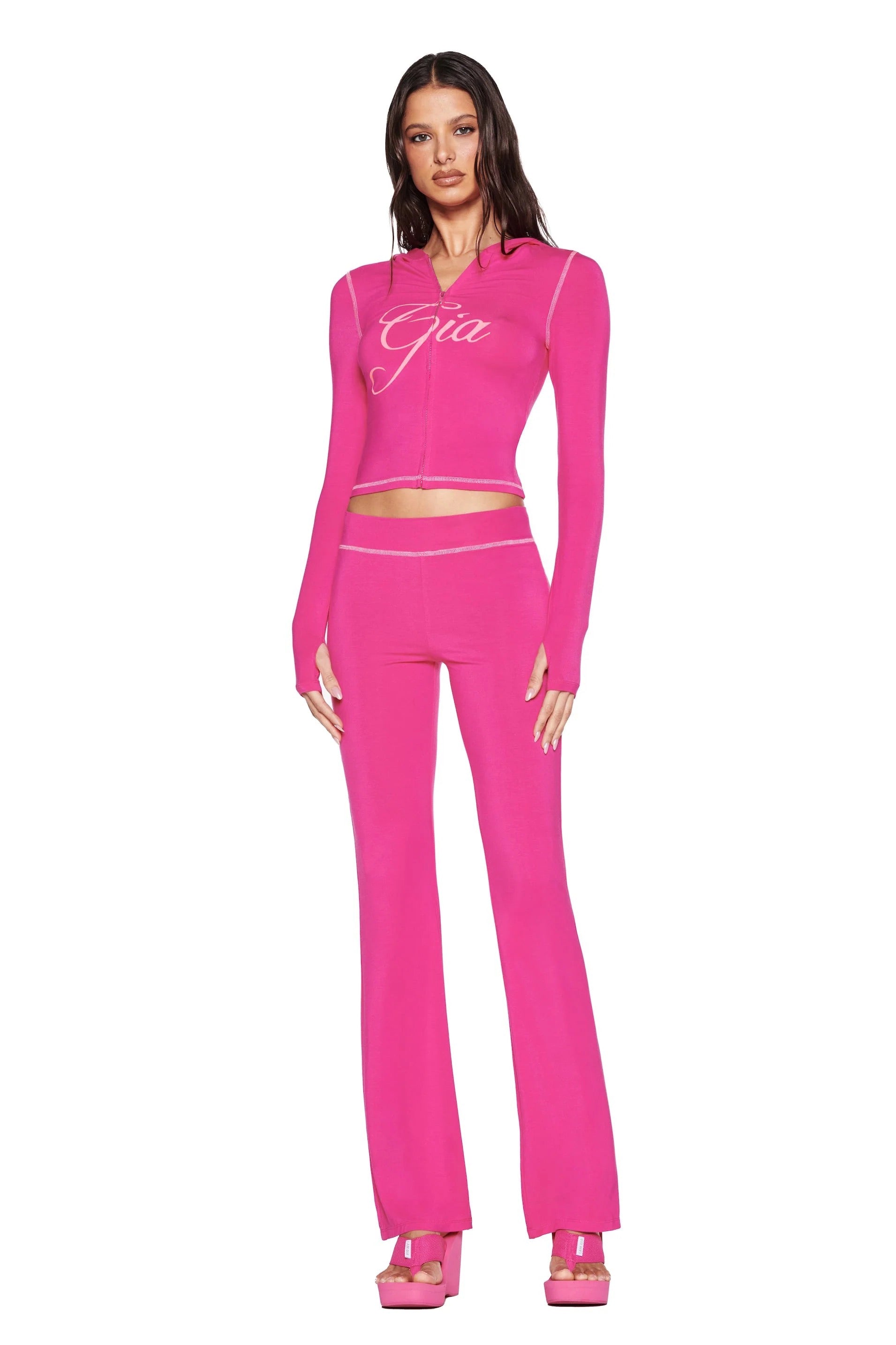 A woman faces forward in I.AM.GIA’s BLARE TRACKPANT - HOT PINK and a matching pink crop top with “Gia,” styled with platform heels against a plain white background.
