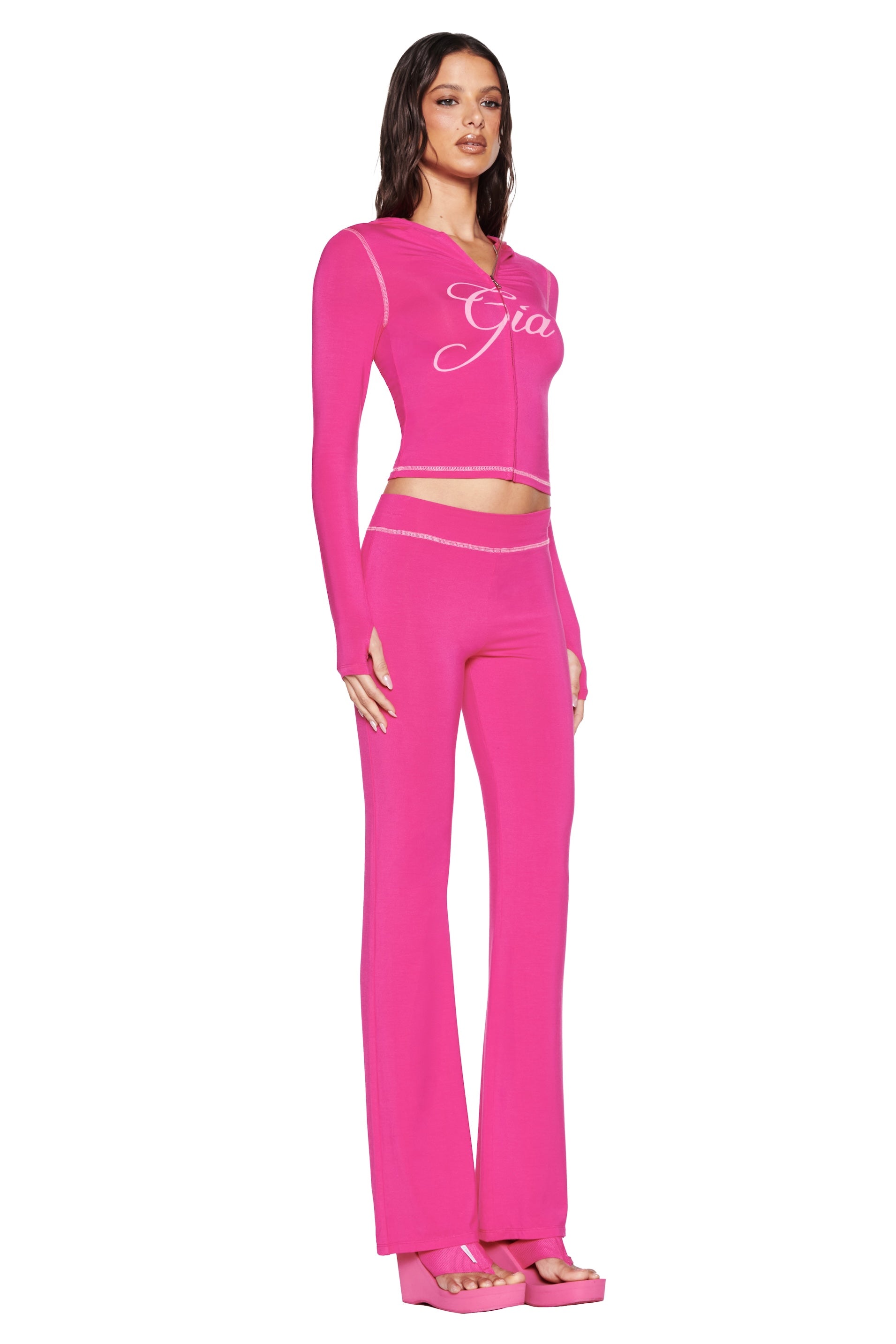 A person in a side pose wears the BLARE TRACKPANT - HOT PINK set from I.AM.GIA, featuring a long-sleeve logo top and trackpants. Their look is completed with pink platform sandals and long, dark hair flowing down their back.