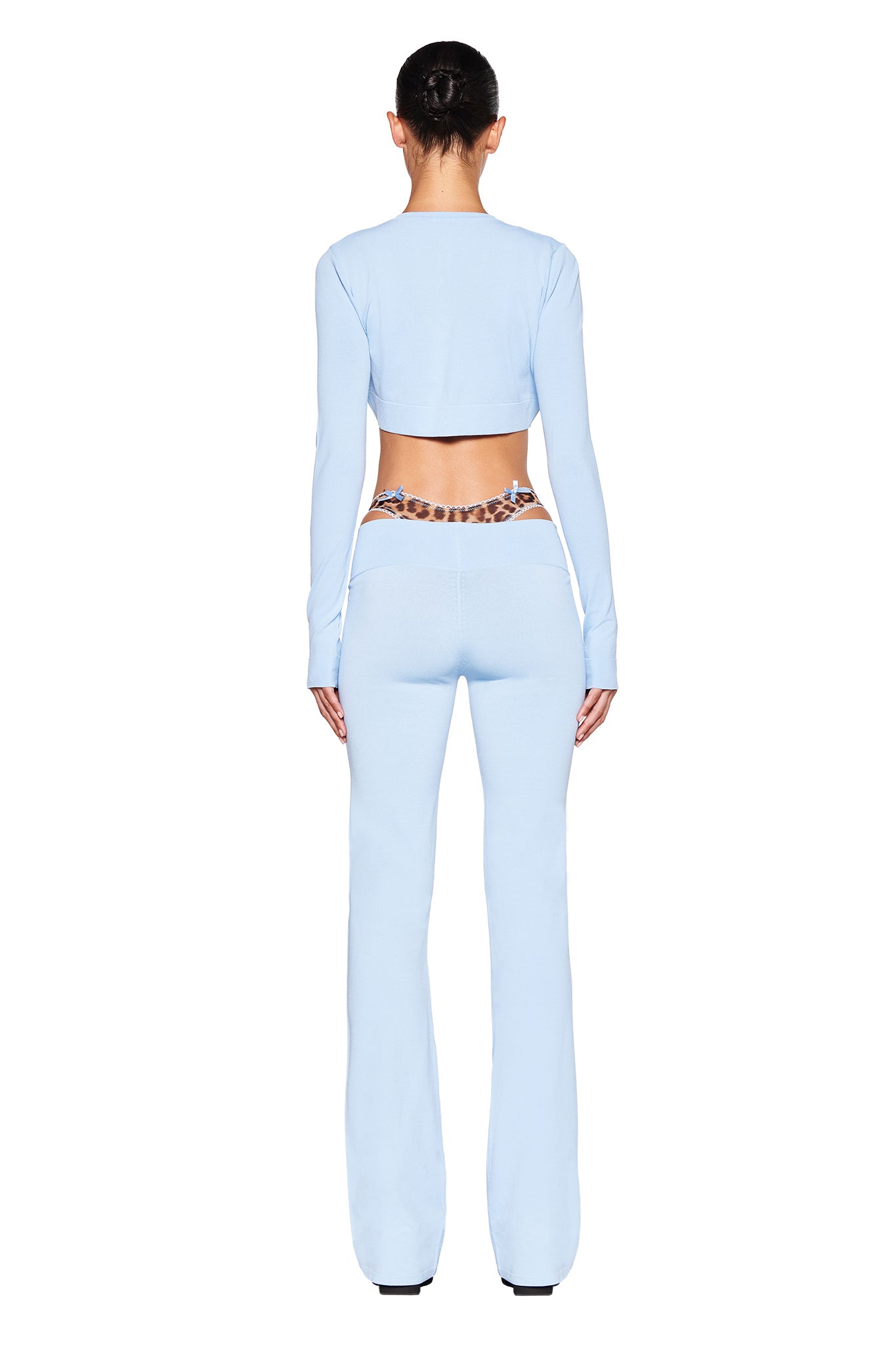 A woman with dark hair in a bun is seen from the back wearing a light blue, long-sleeve crop top and the AUDRINA PANT - BLUE : POWDER BLUE by I.AM.GIA. She has a leopard-print belt around her waist, and the background is plain white.