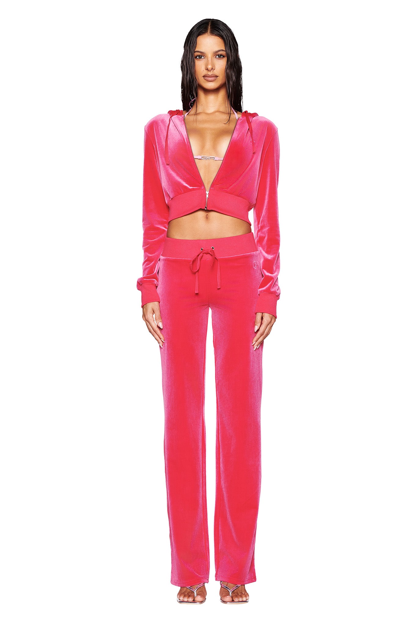 The individual is dressed in a vibrant hot pink STACEY VELOUR PANT from I.AM.GIA, featuring a luxurious velour finish that exudes a shiny texture. The ensemble includes custom 'G' embroidery on both the cropped hoodie and drawstring pants, set against a simple white background.