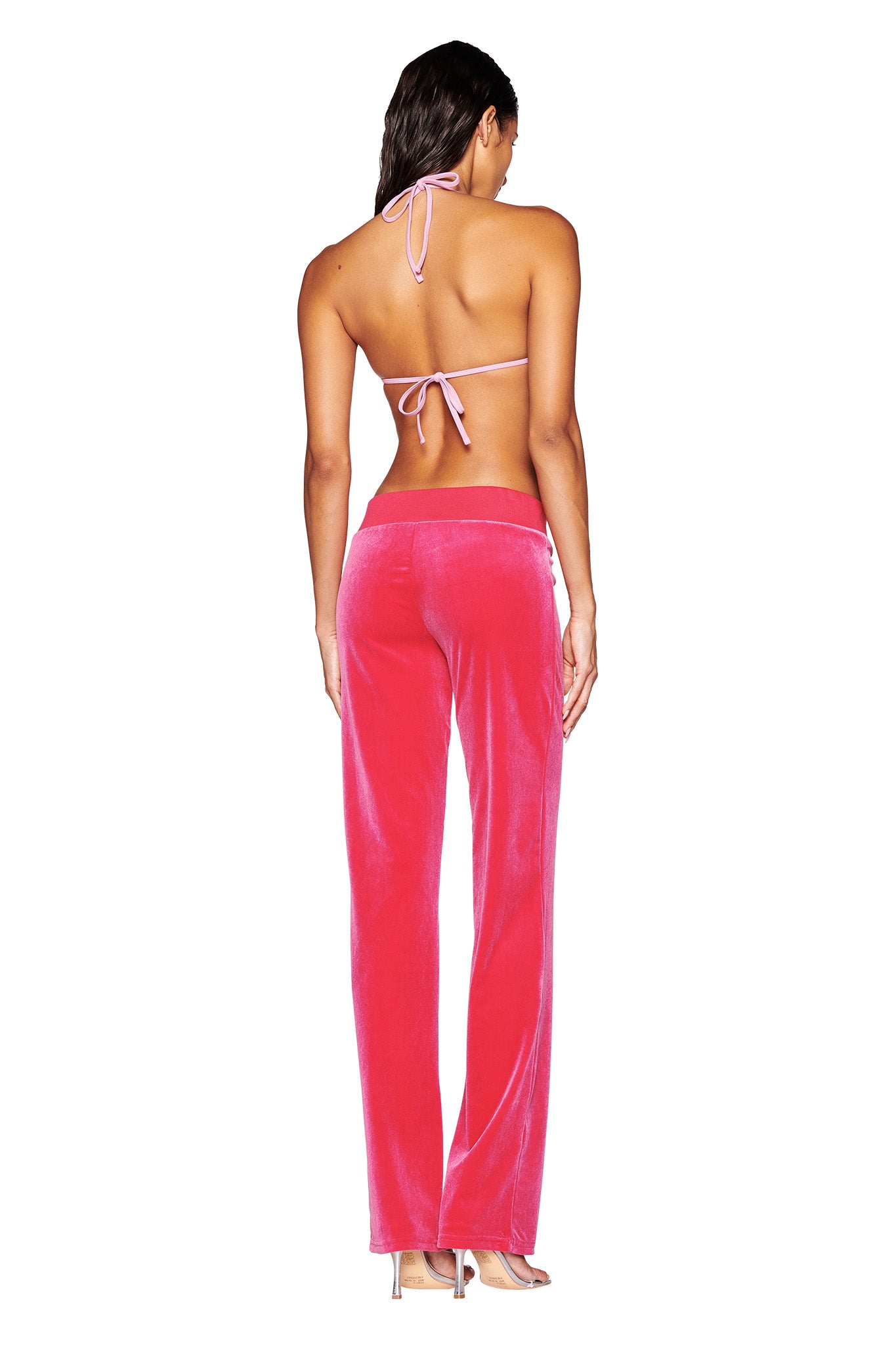 A woman with long hair is depicted from the back, wearing a pink halter bikini top and the HOT PINK STACEY VELOUR PANT by I.AM.GIA, which features a mid-rise waist. She is standing in high-heeled shoes against a white background.
