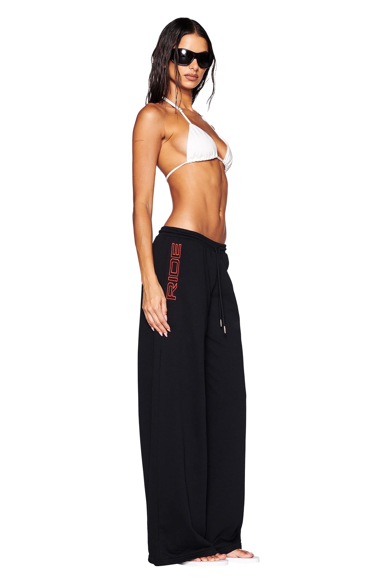 A woman with long dark hair, wearing sunglasses, a white bikini top, and the I.AM.GIA JUSTINE PANT in black featuring GIA embroidery in red lettering, stands confidently barefoot against a white background in profile view.