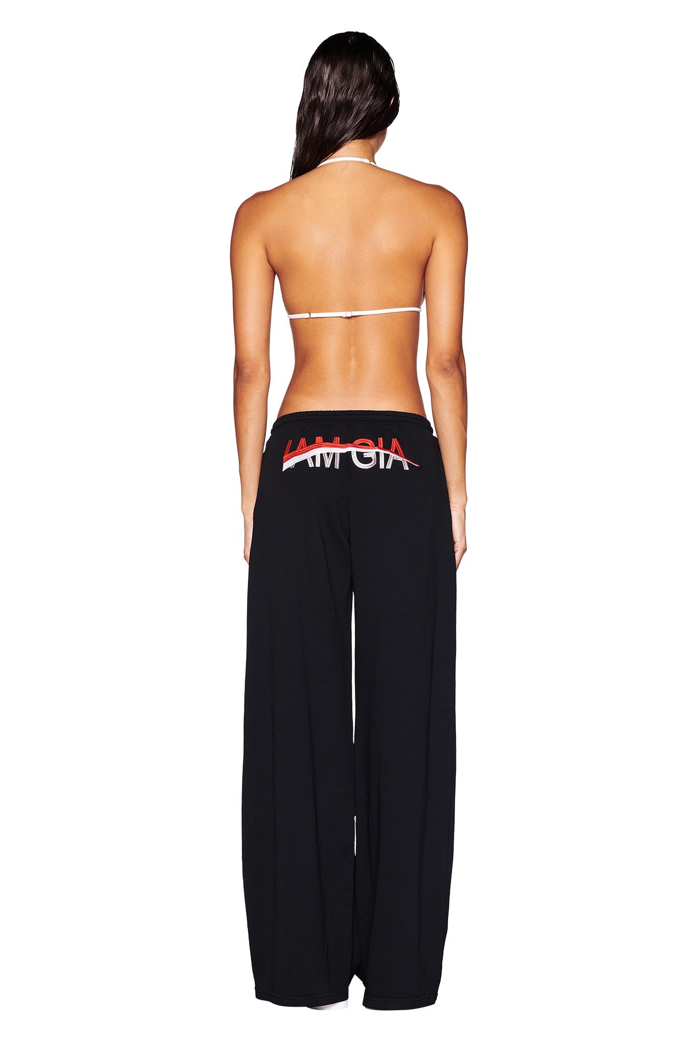 A woman with long brown hair, seen from behind, showcases the I.AM.GIA JUSTINE PANT in black. She pairs a white bikini top with these baggy track pants, which feature GIA embroidery and red and white text on the back. The low-rise waist enhances their casual elegance, as she stands against a plain white background.