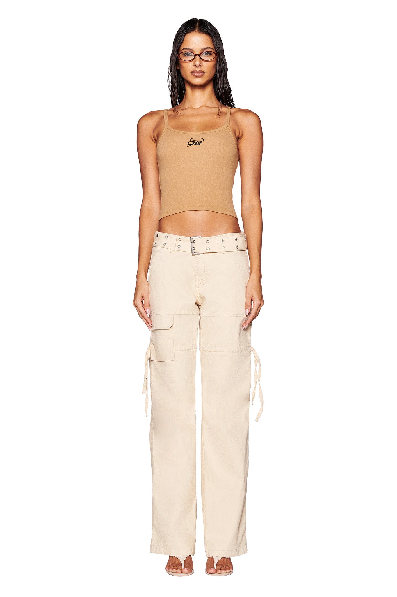 A person stands against a white background, wearing a tan sleeveless crop top, the I.AM.GIA Naima Cargo Pant - Tan with tied side pockets and a low-rise waist, and heels. They have long hair, glasses, a neutral expression, and are accessorized with a necklace.