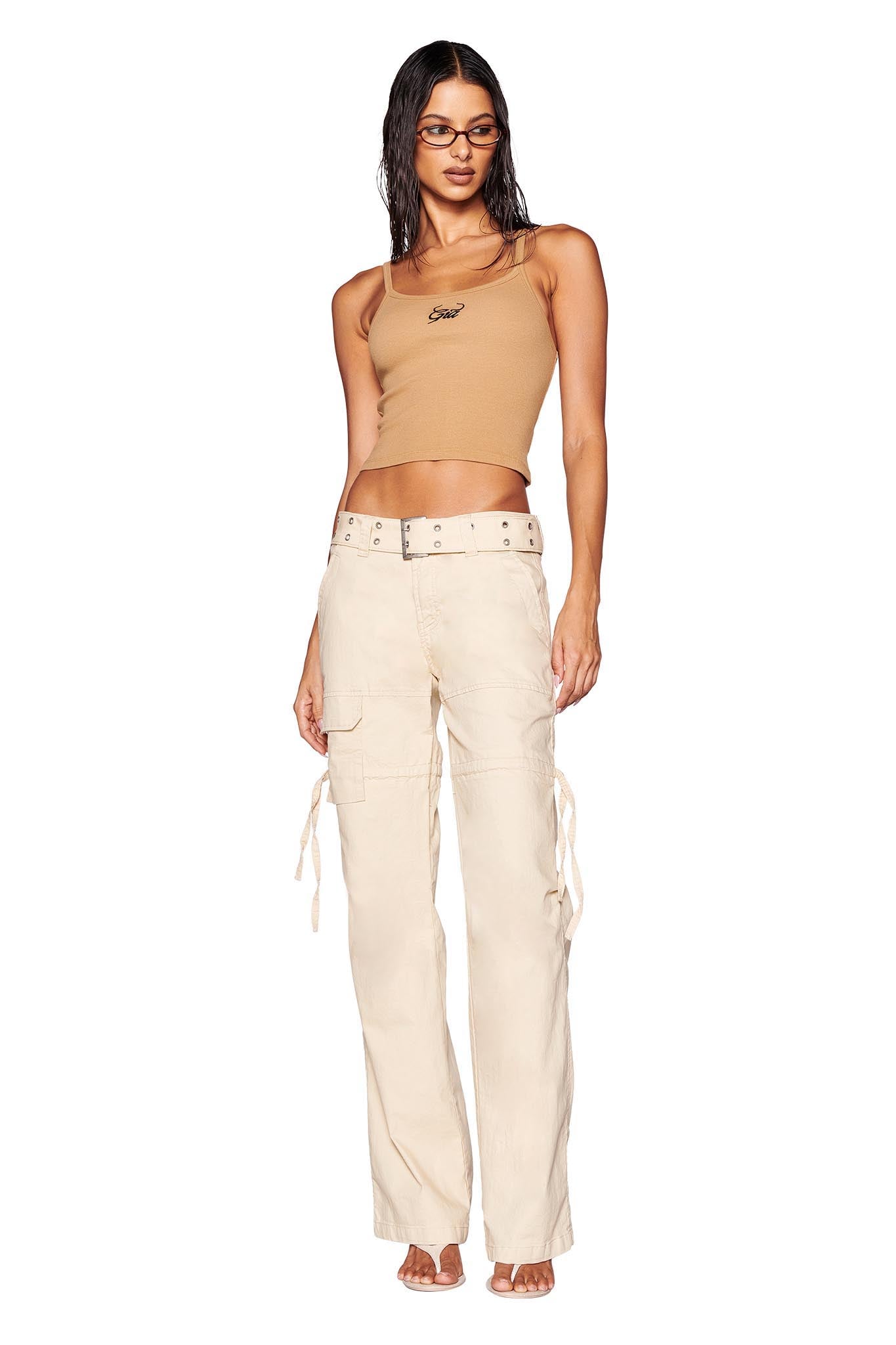 A person with long hair stands confidently in a sleeveless brown crop top and the Naima Cargo Pant from I.AM.GIA in tan, featuring a low-rise waist. They are wearing glasses and light-colored open-toe shoes, accessorized with a belt and a necklace. The background is solid white.
