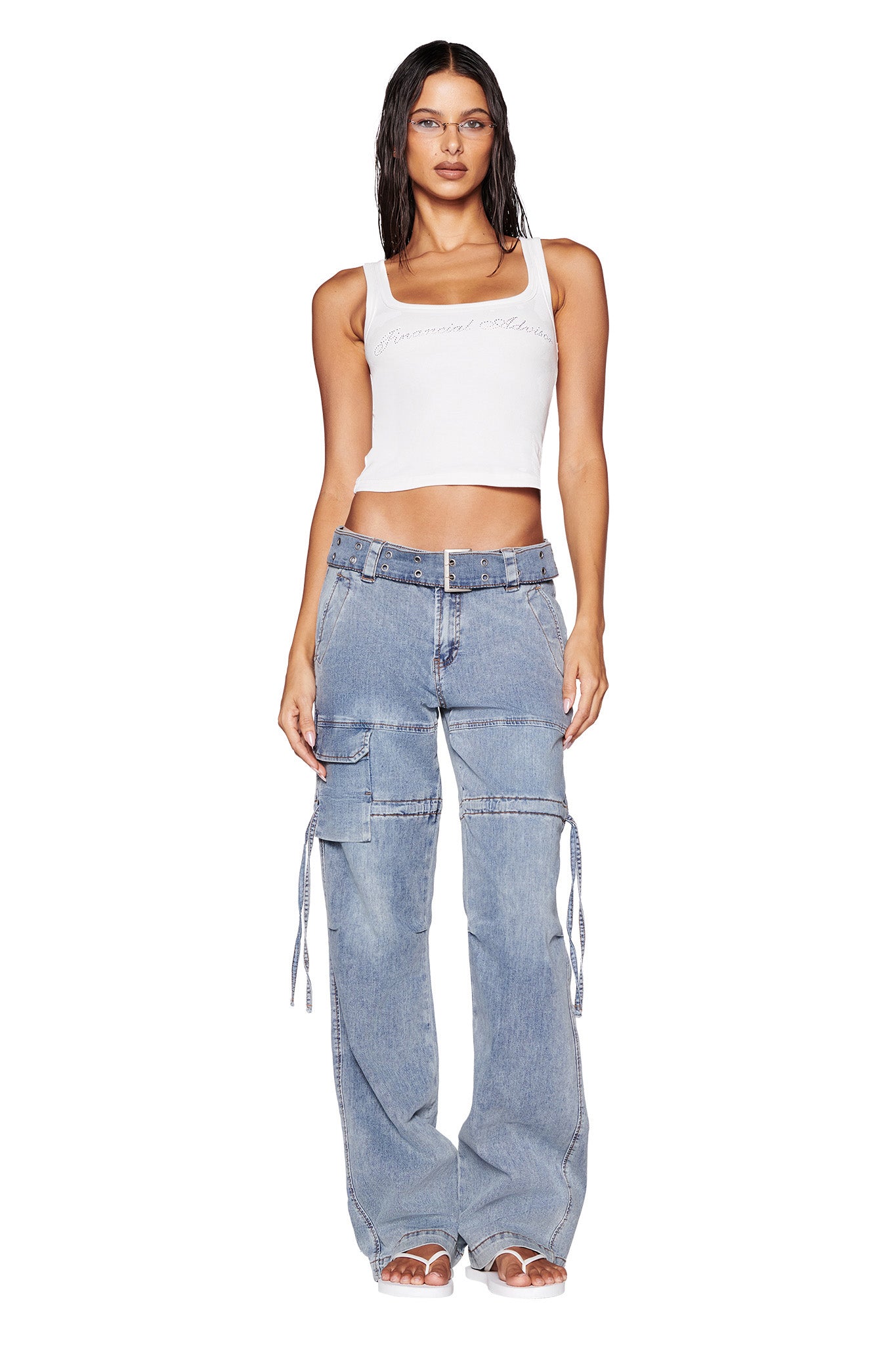 A person with wet, dark hair stands against a white background, wearing a white sleeveless crop top and the Naima Cargo Pant made from 100% cotton by I.AM.GIA. The light blue denim pants feature multiple pockets and straps and are designed with a low-rise waist. They complete the look with white shoes.