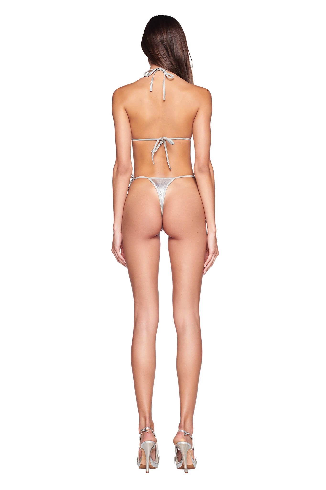 A woman stands with her back to the camera, wearing a silver bikini and high-heeled sandals by I.AM.GIA. She has long, straight hair cascading down her back. The bikini features a halter top with ties at the neck and back, and she is wearing the ALOHI BIKINI BOTTOM - GREY : SILVER in a thong-style.