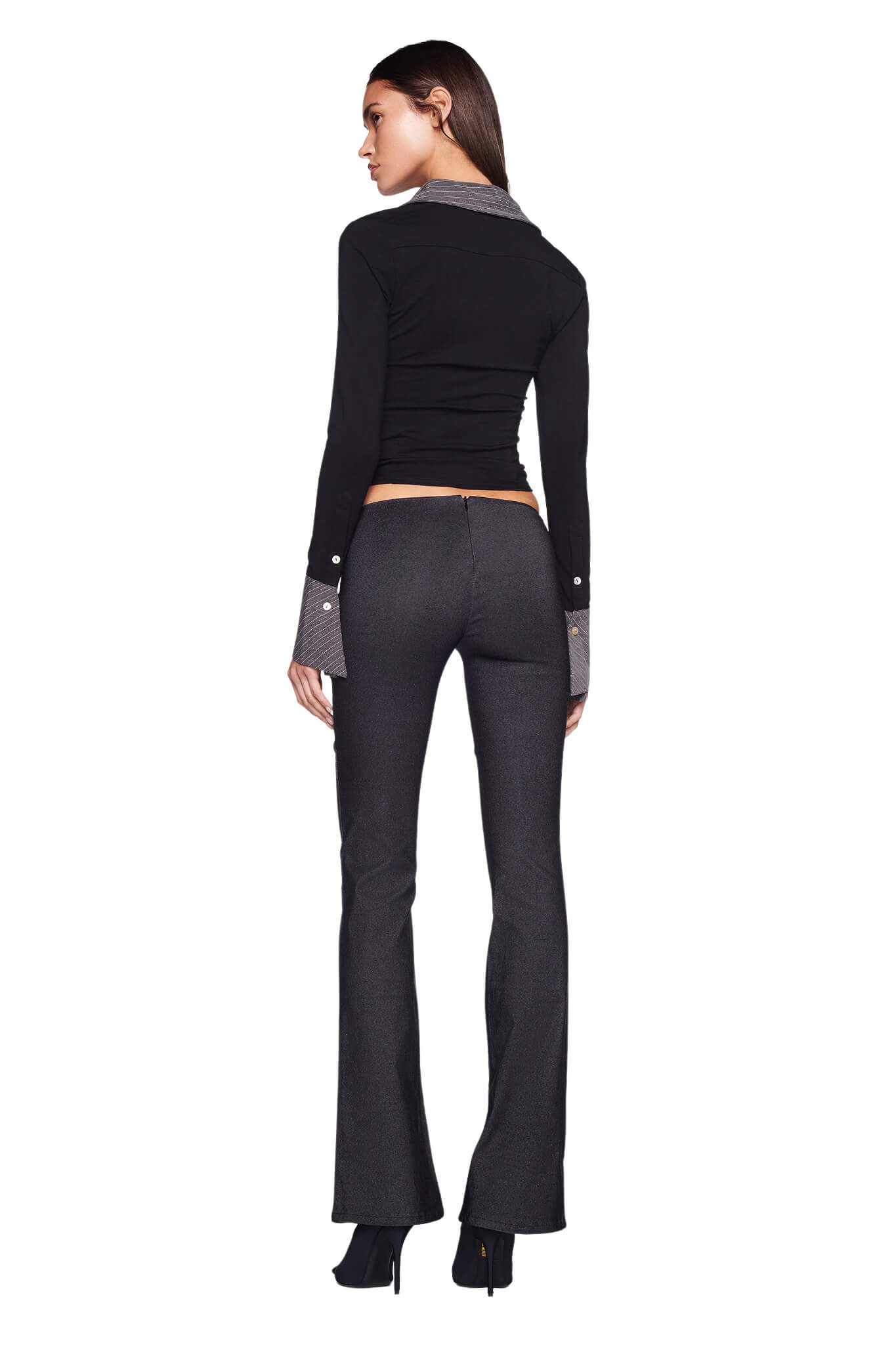 A woman with long dark hair is posing with her back to the camera, showcasing her chic I.AM.GIA SHARAH TOP in black. She pairs it perfectly with high-waisted, flared pants in grey and completes the stylish look with high-heeled shoes against a white background.