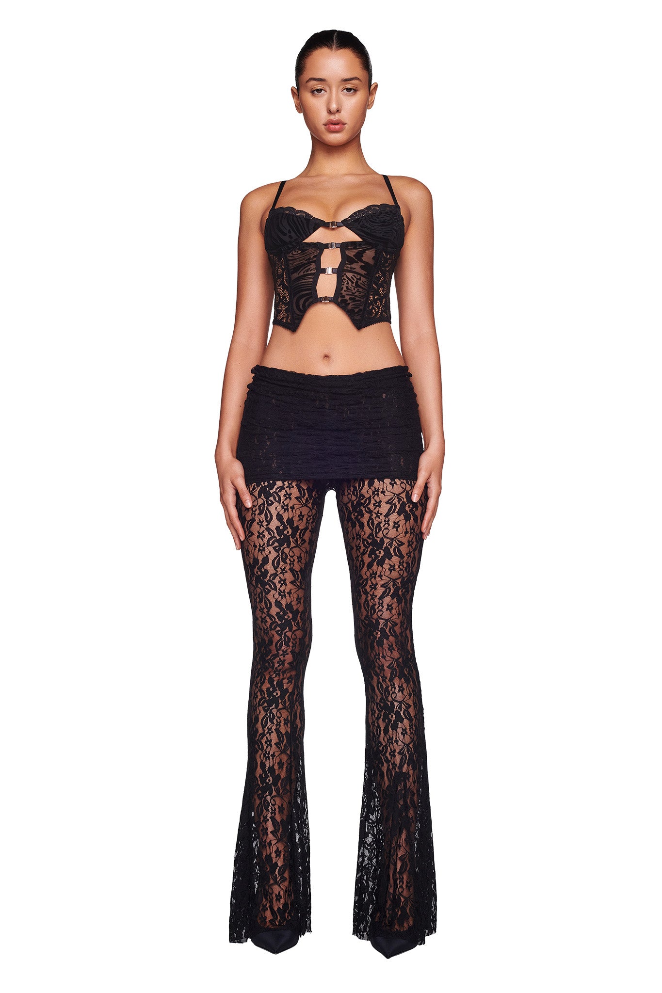 A woman stands against a plain white background, showcasing the I.AM.GIA ELENA TOP - BLACK BURNOUT. She wears a spaghetti strap crop top with cut-out details, paired with an edgy mini skirt and bell-bottom pants. The lace fabric of the pants reveals her legs, adding a bold touch to the ensemble.