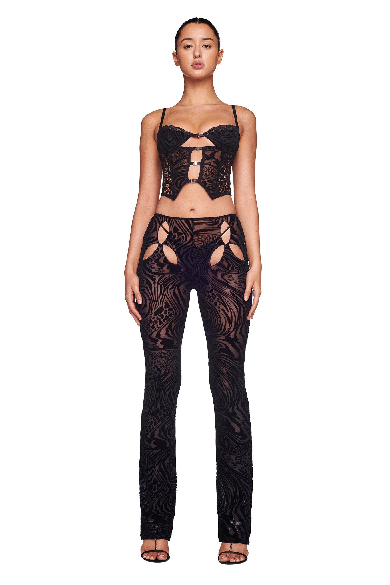 A woman stands against a white background wearing the stylish, form-fitting ELENA TOP - BLACK BURNOUT by I.AM.GIA, paired with matching flared pants featuring cut-out details. She has her hair pulled back and is wearing black heels.