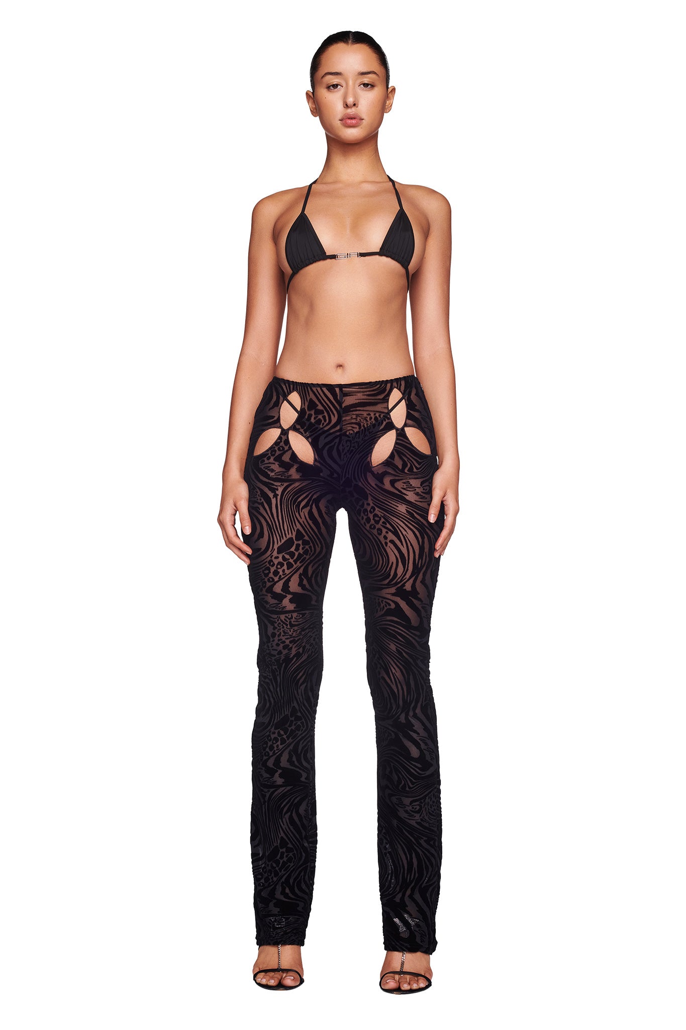 A woman stands facing forward, showcasing the I.AM.GIA AMINA TOP in black burnout and high-waisted patterned black pants with cut-out details at the hips, paired with black heels. Her hair is pulled back, and she displays a neutral expression against a plain white background.