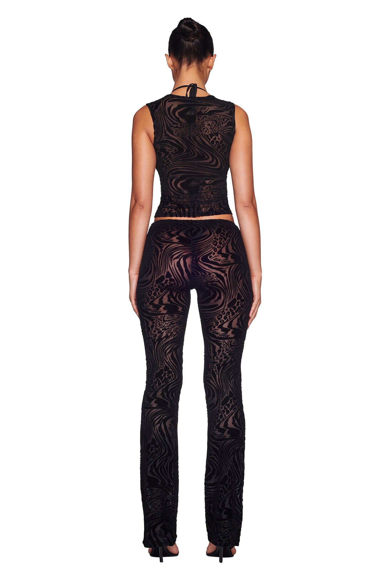 A woman stands facing away from the camera wearing the I.AM.GIA AMINA TOP - BLACK BURNOUT and matching pants featuring intricate, sheer patterns. Her hair is tied in a high bun, and she is wearing black heeled shoes. The background is plain white.