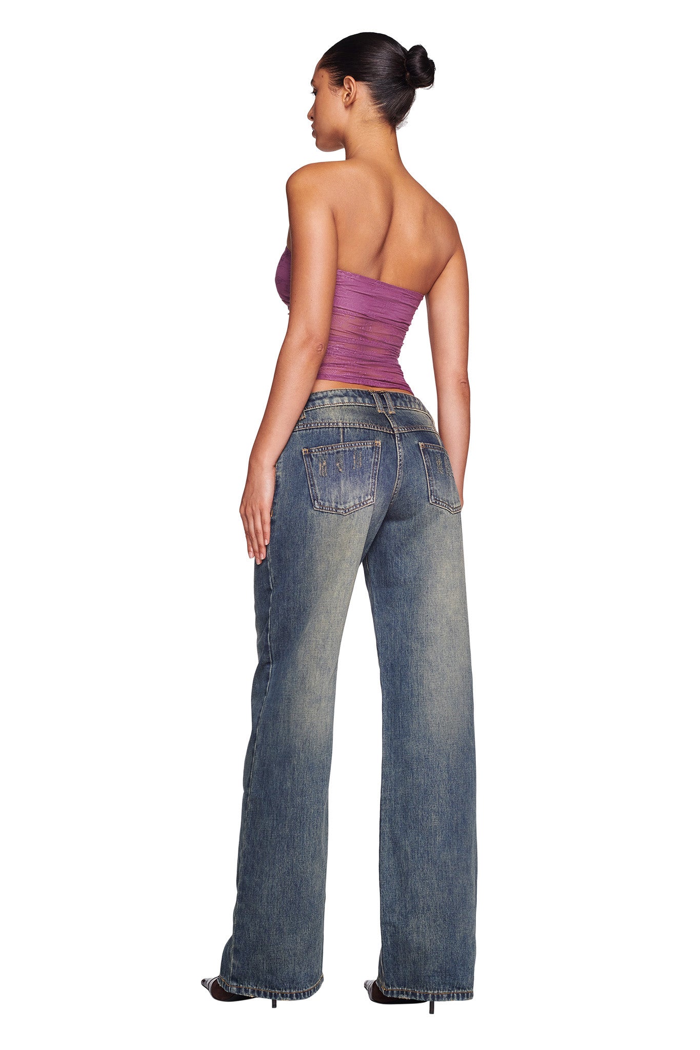 A person stands with their back to the camera, showcasing a strapless, ruched, purple top by I.AM.GIA paired with the DYLAN JEAN in blue denim from I.AM.GIA. The high-waisted, loose-fit jeans feature distressed details and visible pockets. Their hair is styled in a neat bun, highlighting the trendy and casual ensemble.