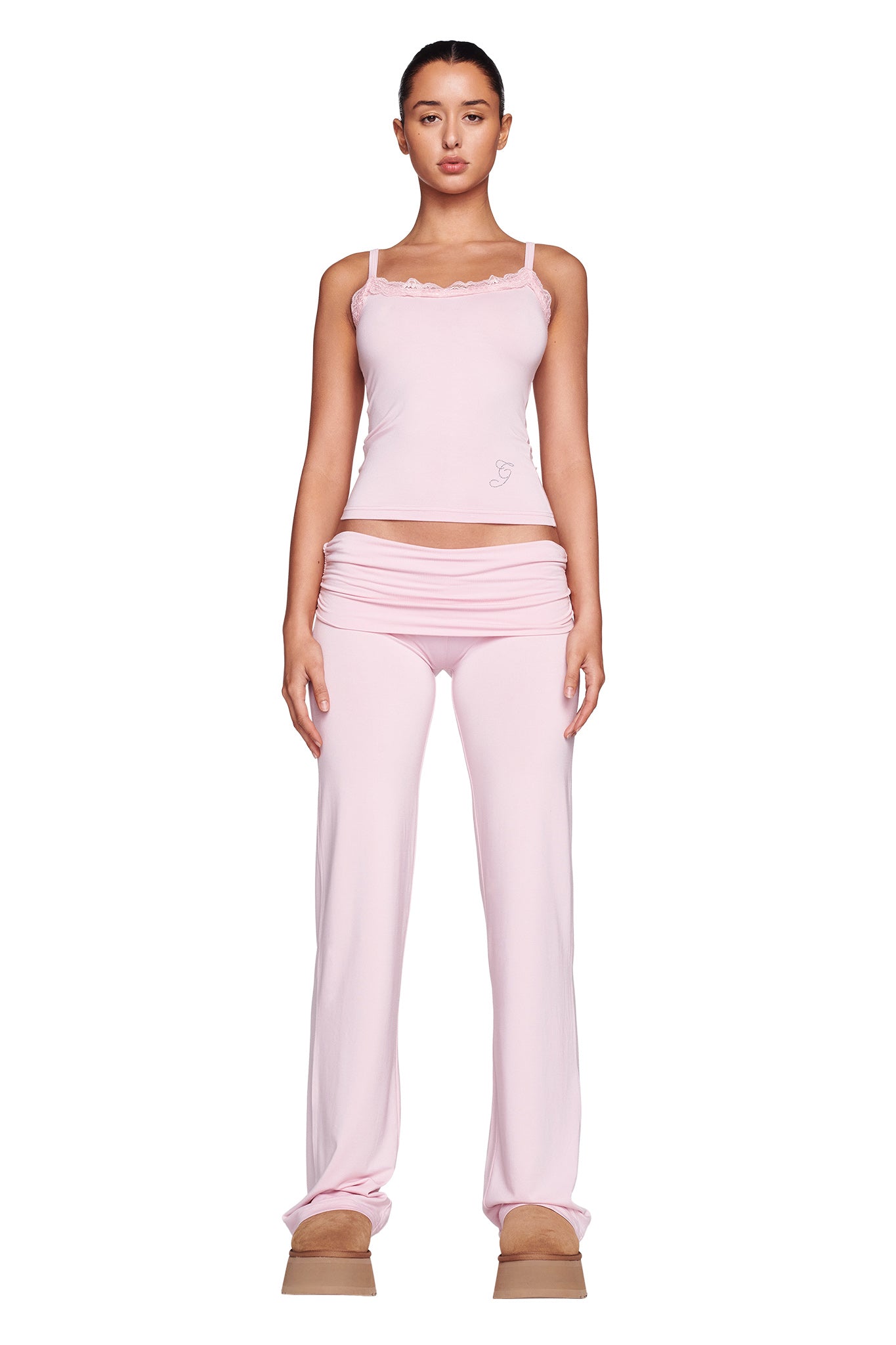A person stands against a white background wearing the I.AM.GIA ERIN TOP in pink, which features a cozy design with lace trim. They have on matching pink low-rise pants and beige platform shoes. Their hair is pulled back, and their arms rest by their sides, highlighting the bodycon silhouette of their ensemble.