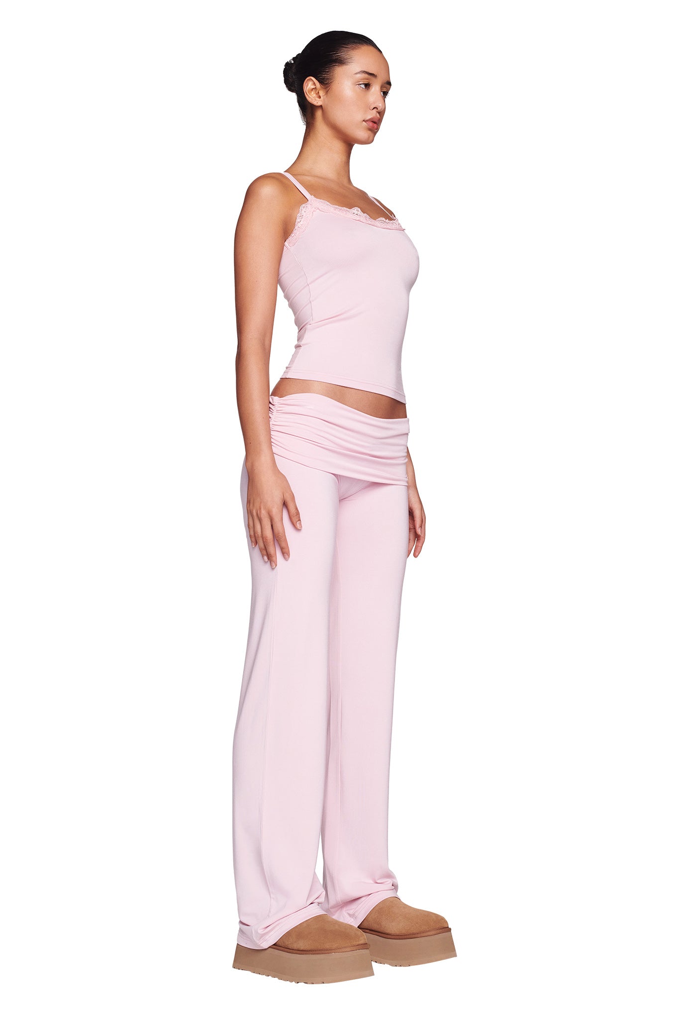 A woman stands sideways, wearing the I.AM.GIA ERIN TOP - PINK, a cozy bamboo-fabric tank top paired with matching loose pants that feature a draped waistband. Her hair is tied back, and she completes the outfit with beige platform shoes. The plain white background accentuates her bodycon silhouette.