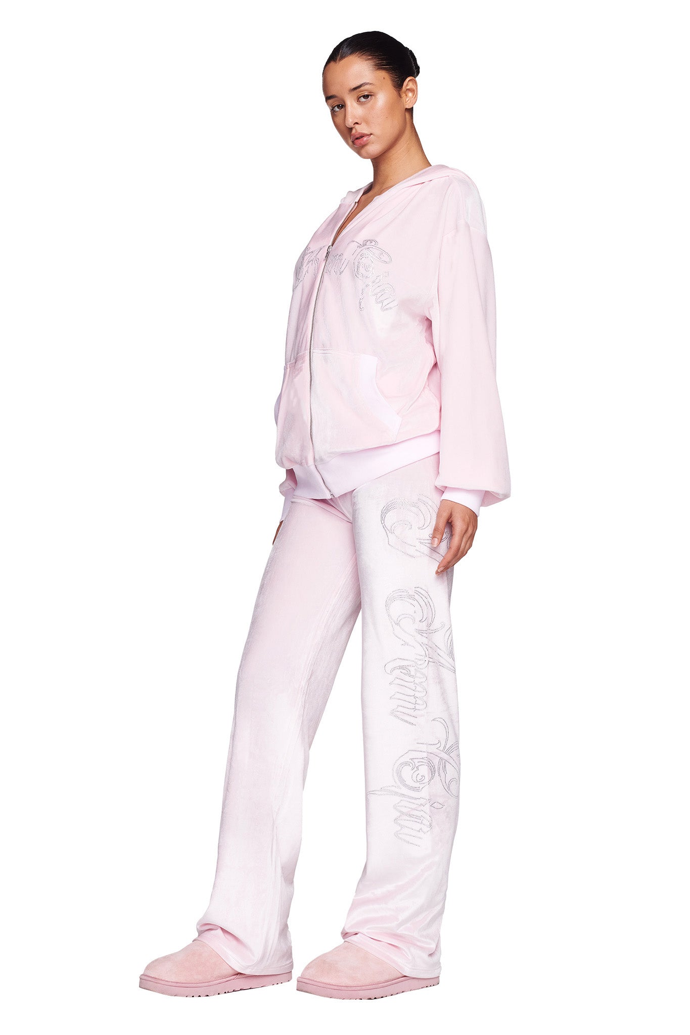 A person in an Icy Pink colourway tracksuit, featuring intricate white embroidery and custom 'I.Am.GIA' logo on the jacket paired with the Daria Pant - Pink : Icy Pink, stands with one leg slightly bent and arms at their sides. Matching slippers complete the look, while dark hair is pulled back with a neutral expression.