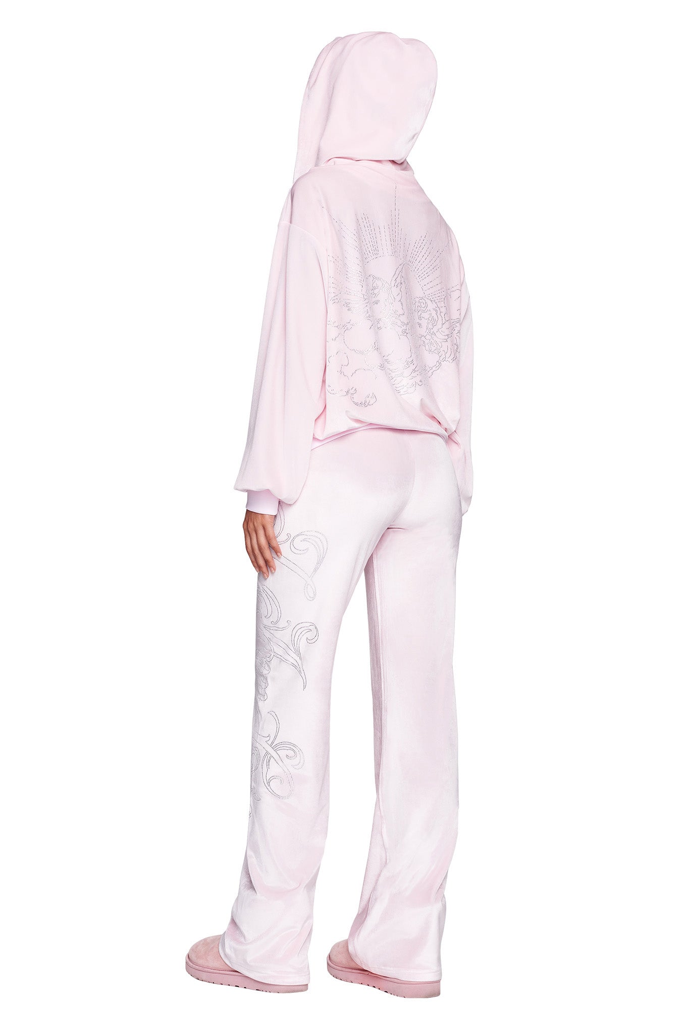 A person is standing with their back facing the camera, dressed in an I.AM.GIA pastel pink DARIA HOODIE – PINK: ICY PINK tracksuit. The outfit boasts intricate designs on the back of the hoodie and along the sides of the pants. They complete their look with matching pink shoes.