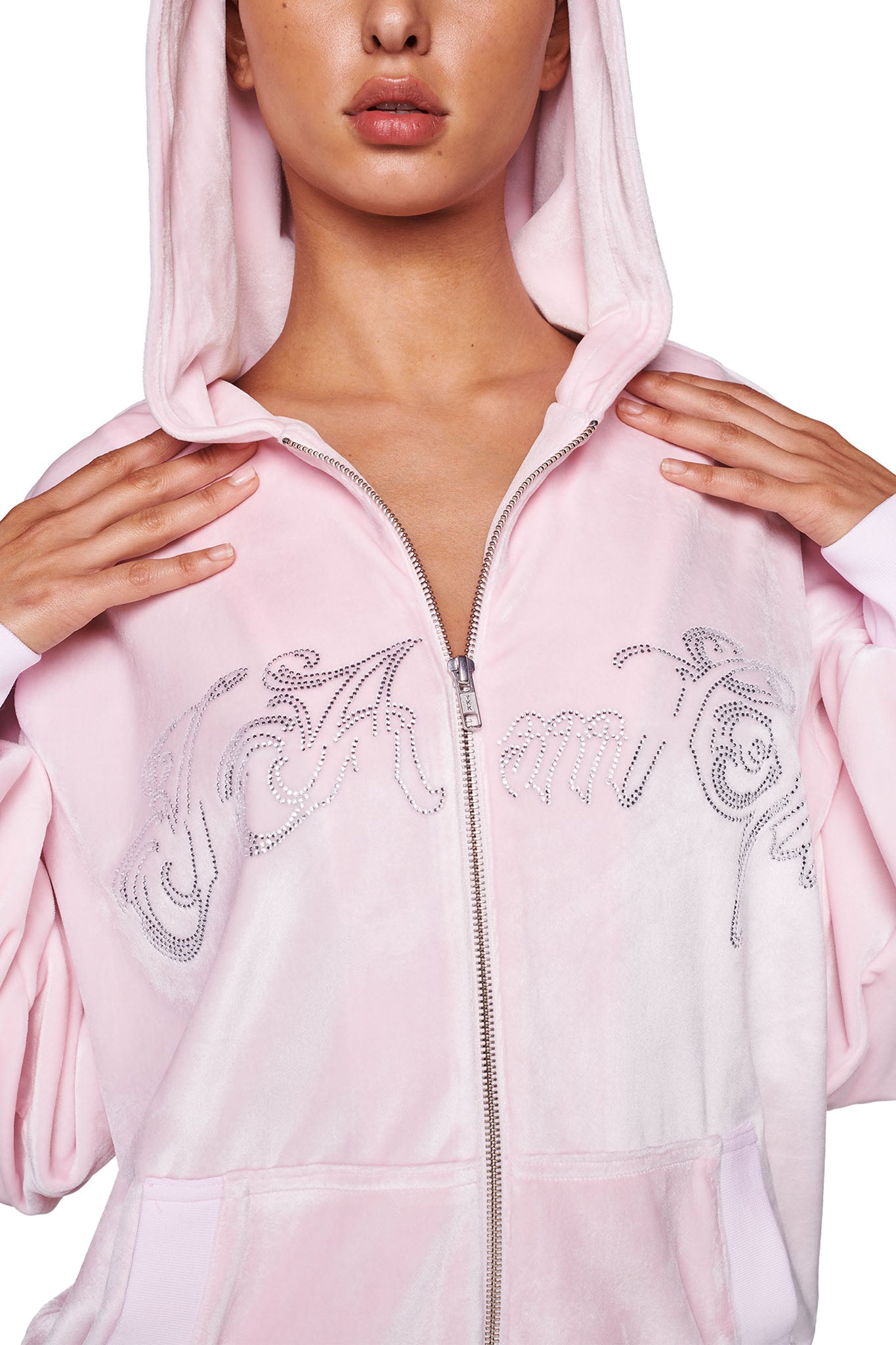 A person wearing an icy pink DARIA HOODIE from I.AM.GIA with the hood up, partially pulled over their head. The hoodie is zipped up and features sparkling rhinestone designs. Only the lower half of their face is visible, and their hands are holding the edges of the hood.