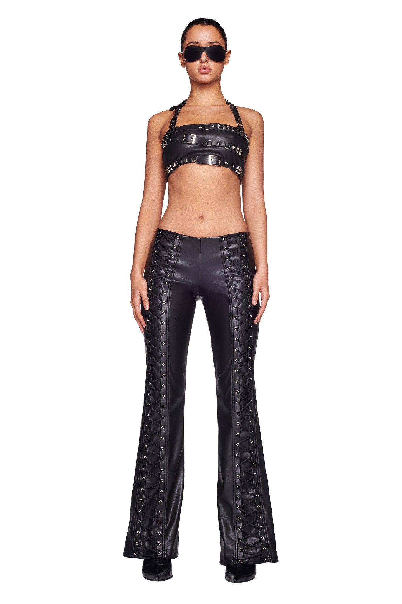 A person stands confidently against a white background, dressed in the SONORA PANT - BLACK from I.AM.GIA. Their ensemble includes high-waisted pants with intricate lace-up detailing and a black leather cropped top adorned with metal studs. They complete the look with black sunglasses and shoes.