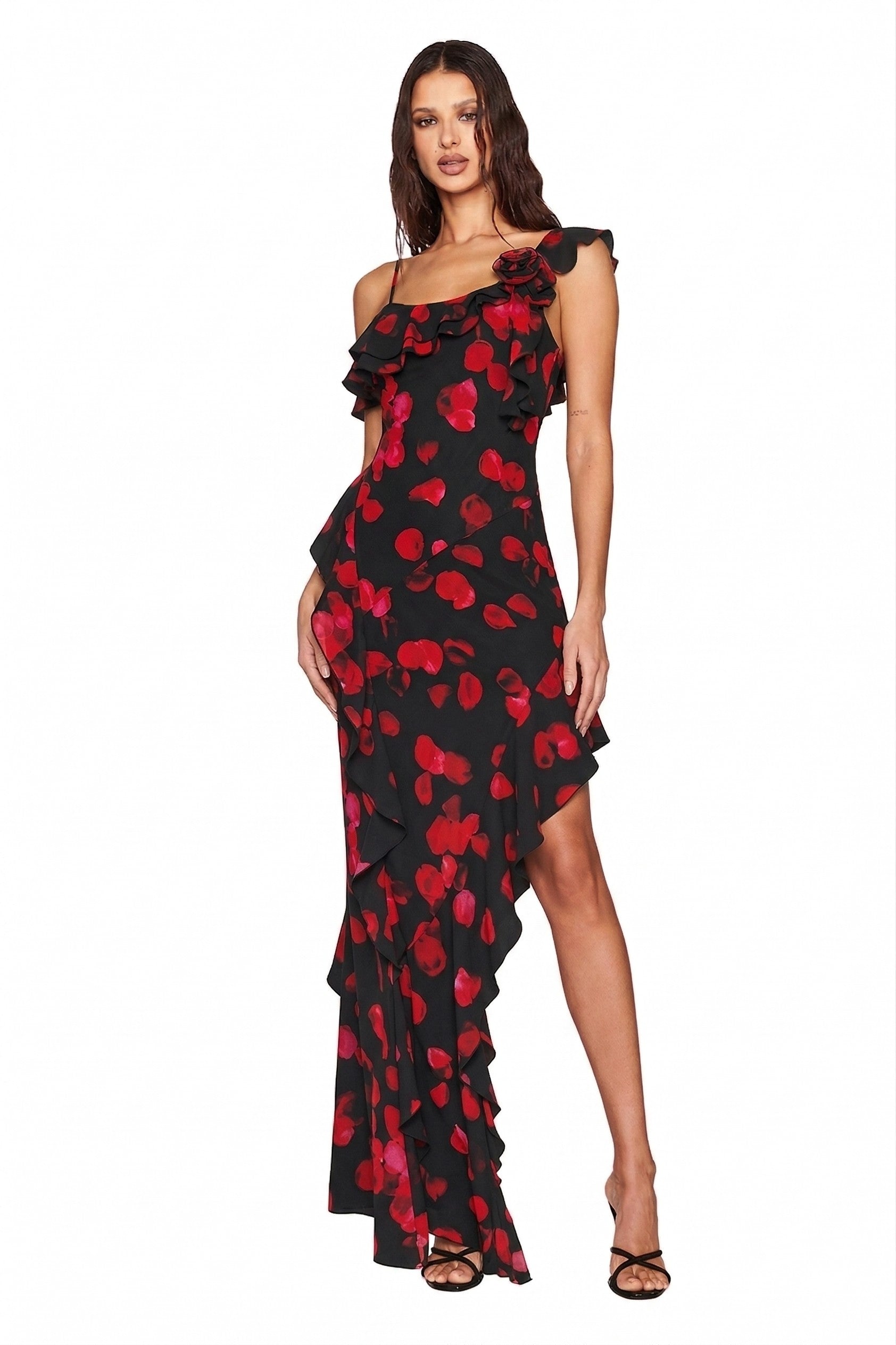 A woman poses against a white background in the I.AM.GIA JULIETTA MAXI DRESS - BLACK PRETTY PETAL, featuring a one-shoulder ruffle and bold red petal print. She stands with one leg forward, hand on her hip.