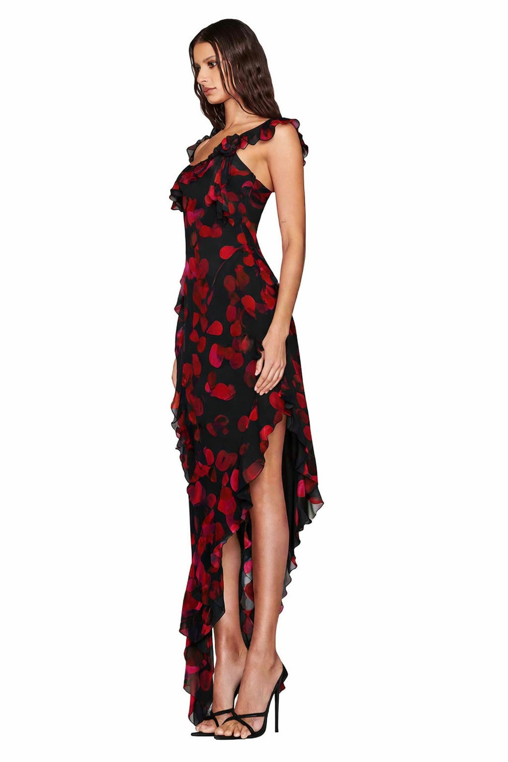 A woman in profile wears the I.AM.GIA JULIETTA MAXI DRESS - BLACK PRETTY PETAL, featuring a rose petal print, one-shoulder ruffle, and asymmetrical high-low hem, paired with high heels against a white background.