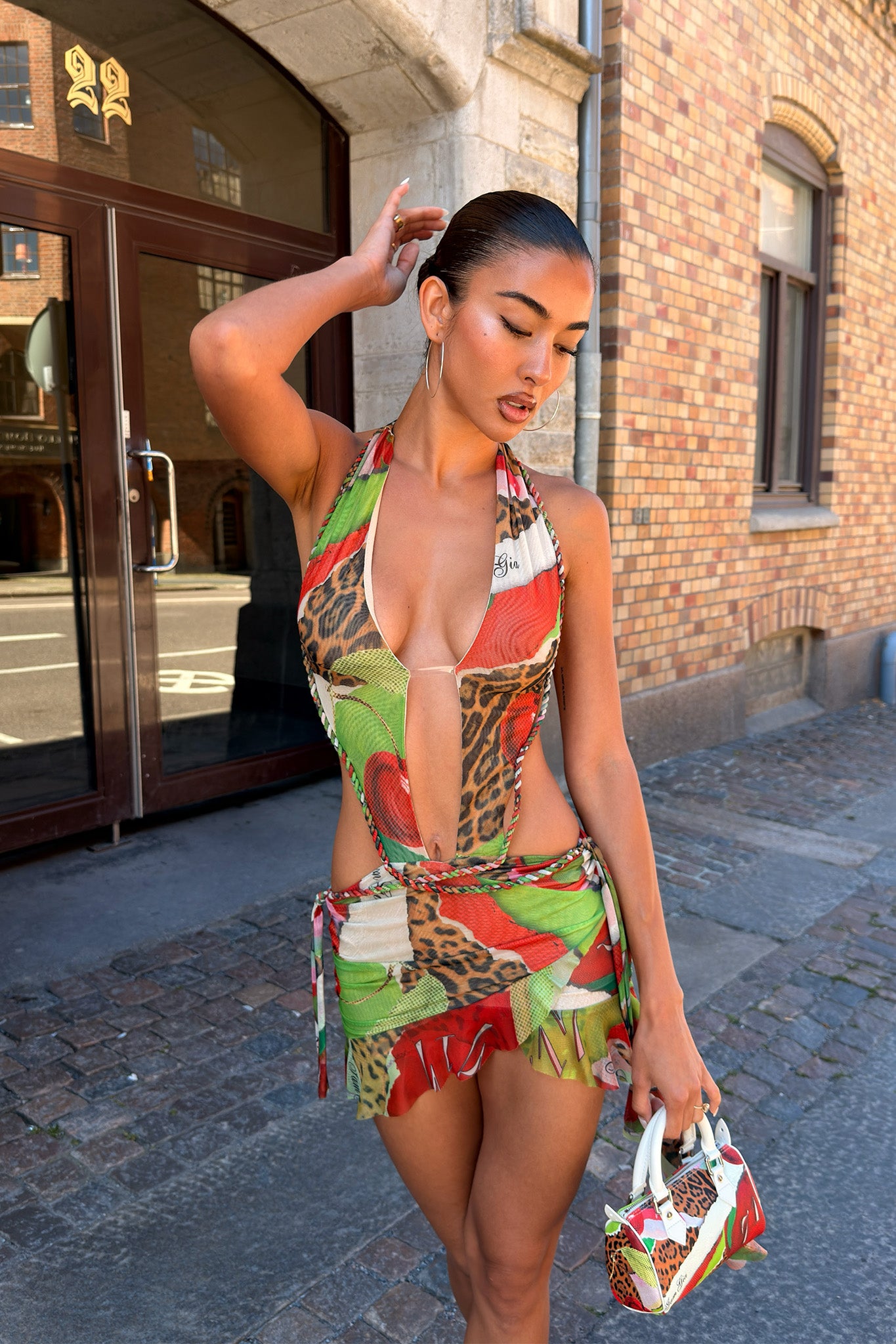 A woman stands on a cobblestone street, confidently posing in the I.AM.GIA AERIN MINI DRESS - WILD CHERRY, a vibrant halter v-neck mini dress with bold animal and cherry prints. She holds a small patterned handbag, one hand touching her head.