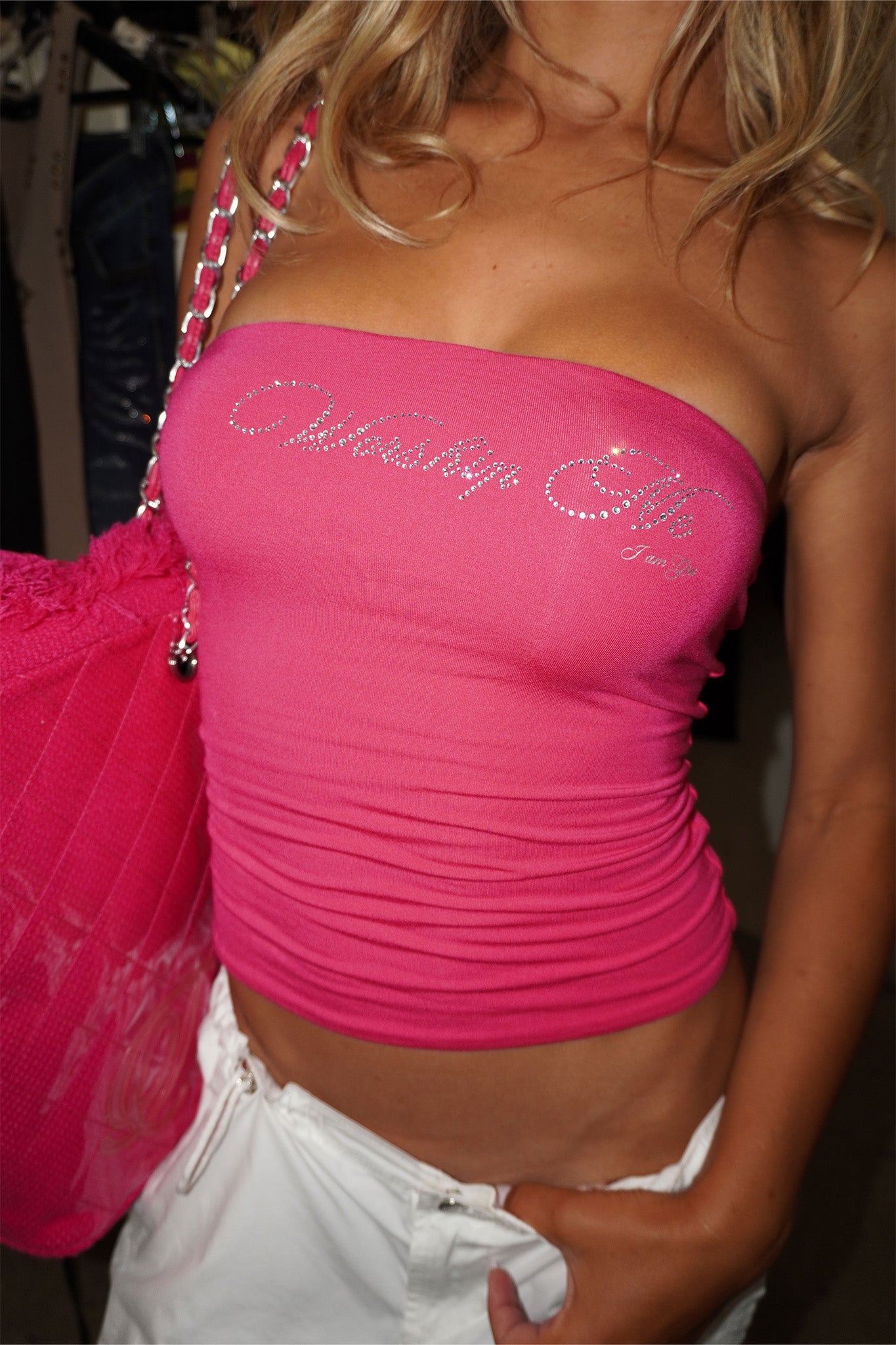 Someone wearing the I.AM.GIA NESSY BOOB TUBE - WORSHIP ME in hot pink with a rhinestone graphic and white shorts holds a matching pink bag. The background features blurred clothing racks.