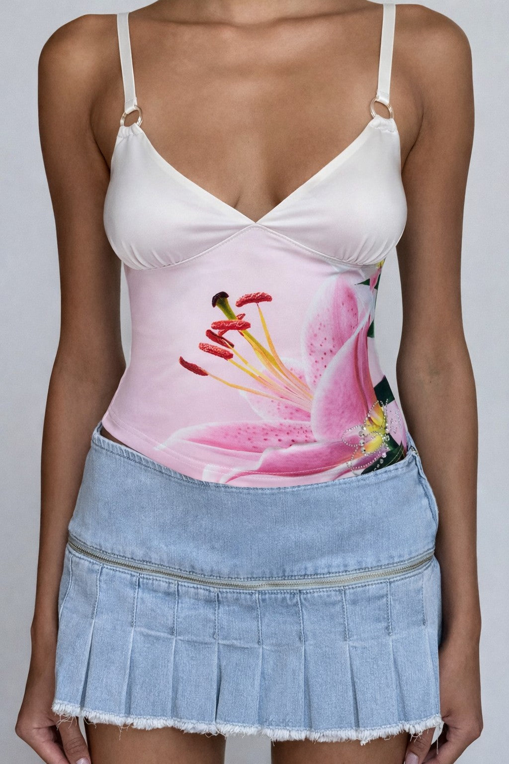 A woman wears the I.AM.GIA ODETTA TOP in Pink Lily Ombre, a pink floral camisole, paired with a light blue pleated denim mini skirt with frayed hem, standing against a plain background.