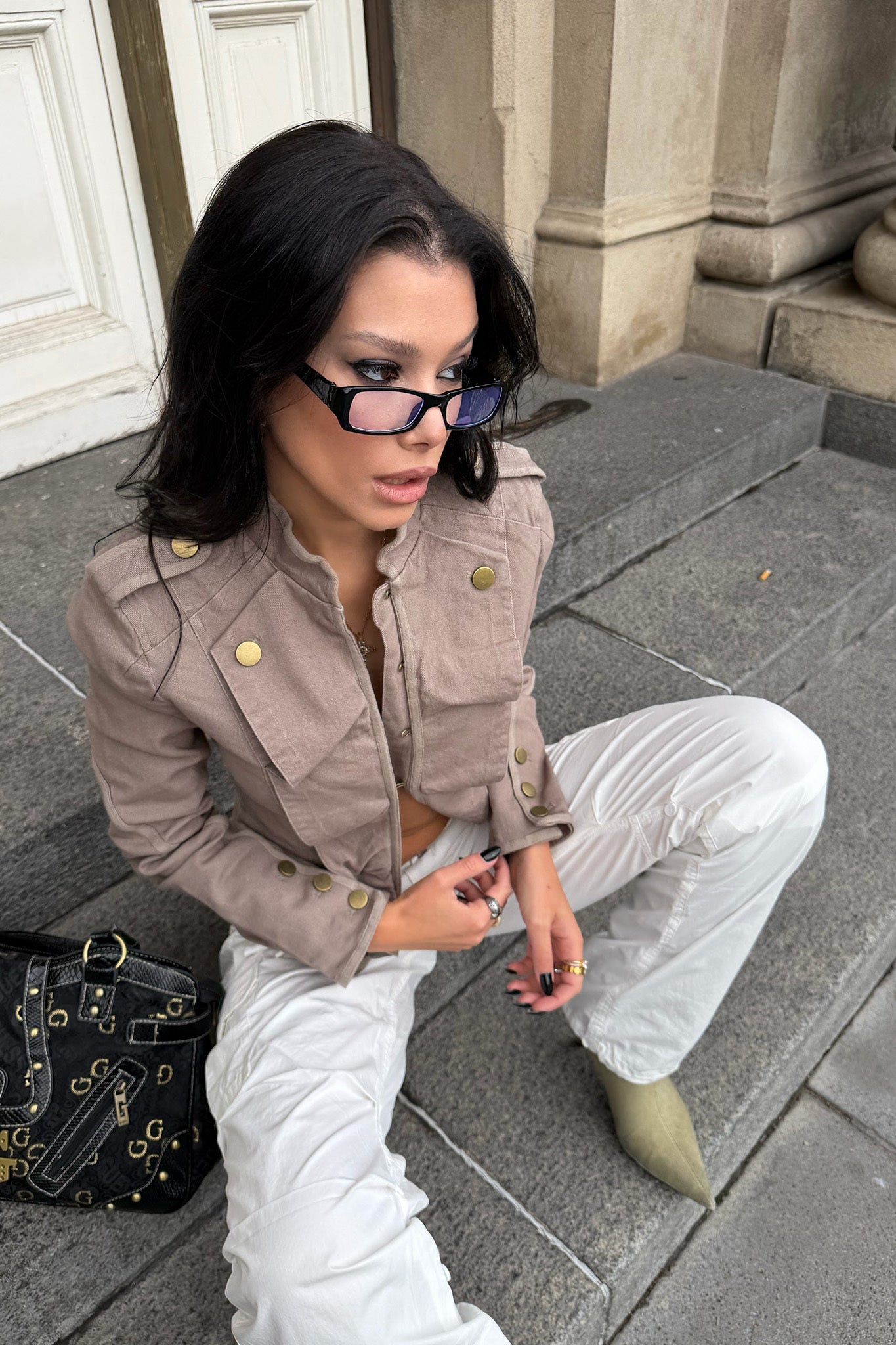 A woman with long dark hair wears the I.AM.GIA SHARN JACKET in Mushroom, white pants, green shoes, blue-framed glasses, and holds sunglasses as she sits on stone steps by a building. A black bag is beside her.