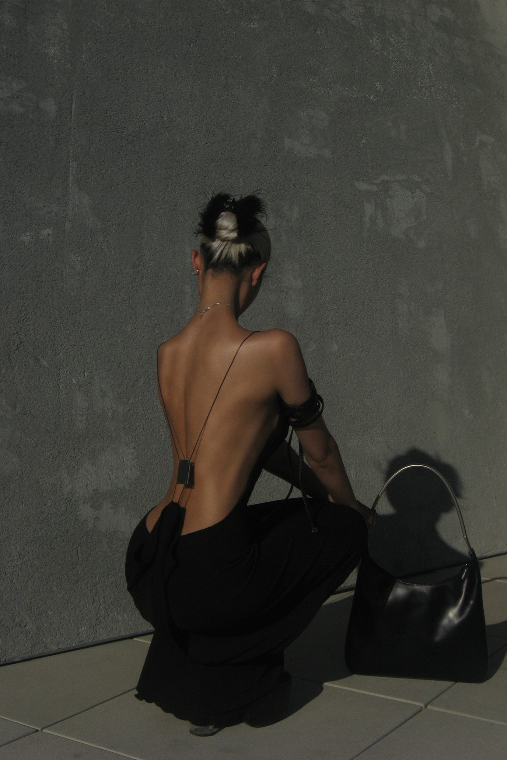 A person with a bleach-blonde and black updo kneels facing a textured gray wall, wearing the I.AM.GIA SIMONE MAXI DRESS in black. A large black handbag sits on the ground beside them.