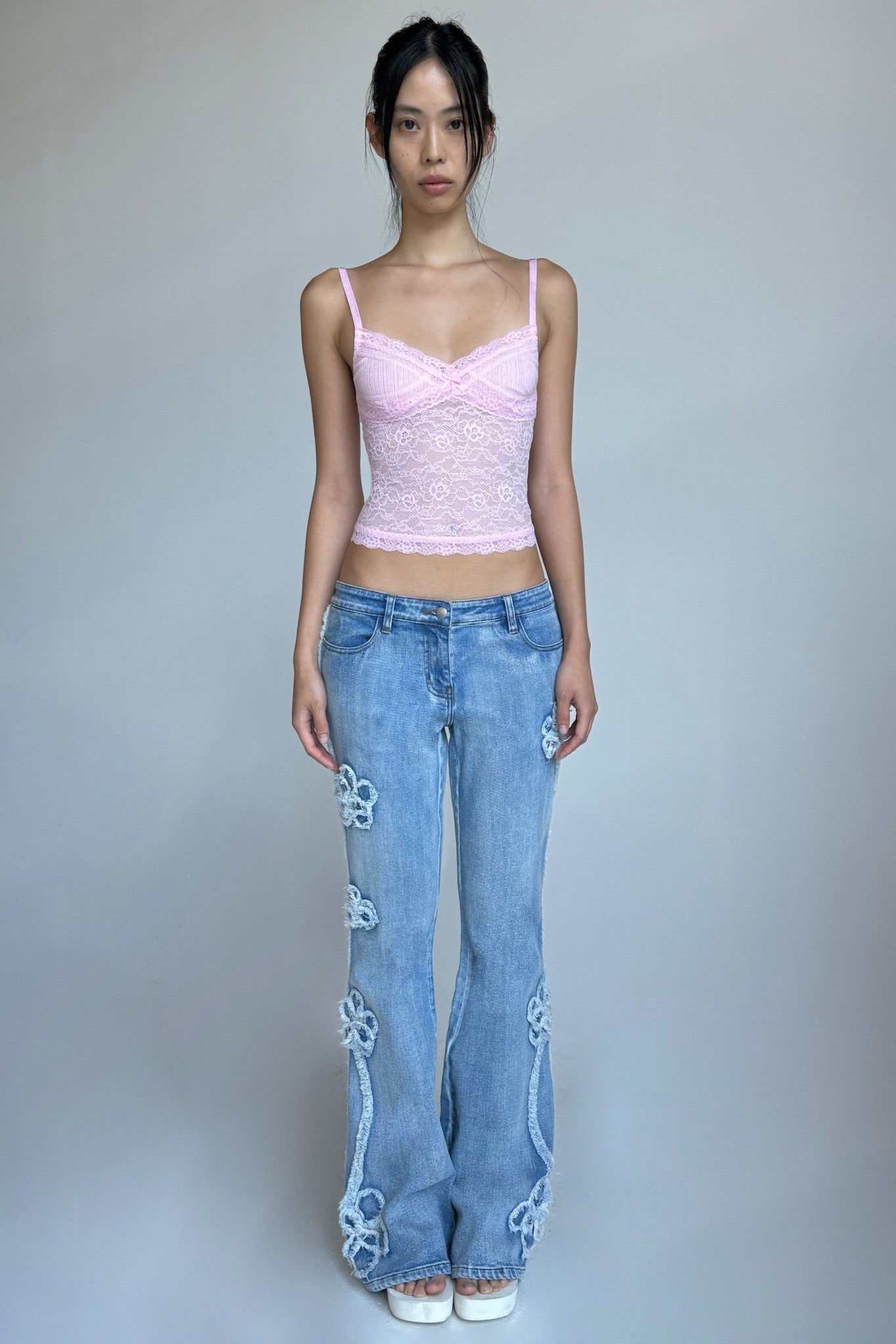 A woman stands against a plain background, wearing a light pink lacy camisole and the TYLA JEAN - BLUE: DENIM BLUE jeans from I.AM.GIA, which are adorned with floral appliqués. She pairs the outfit with white platform shoes. Her hair is tied back and she has a neutral expression.