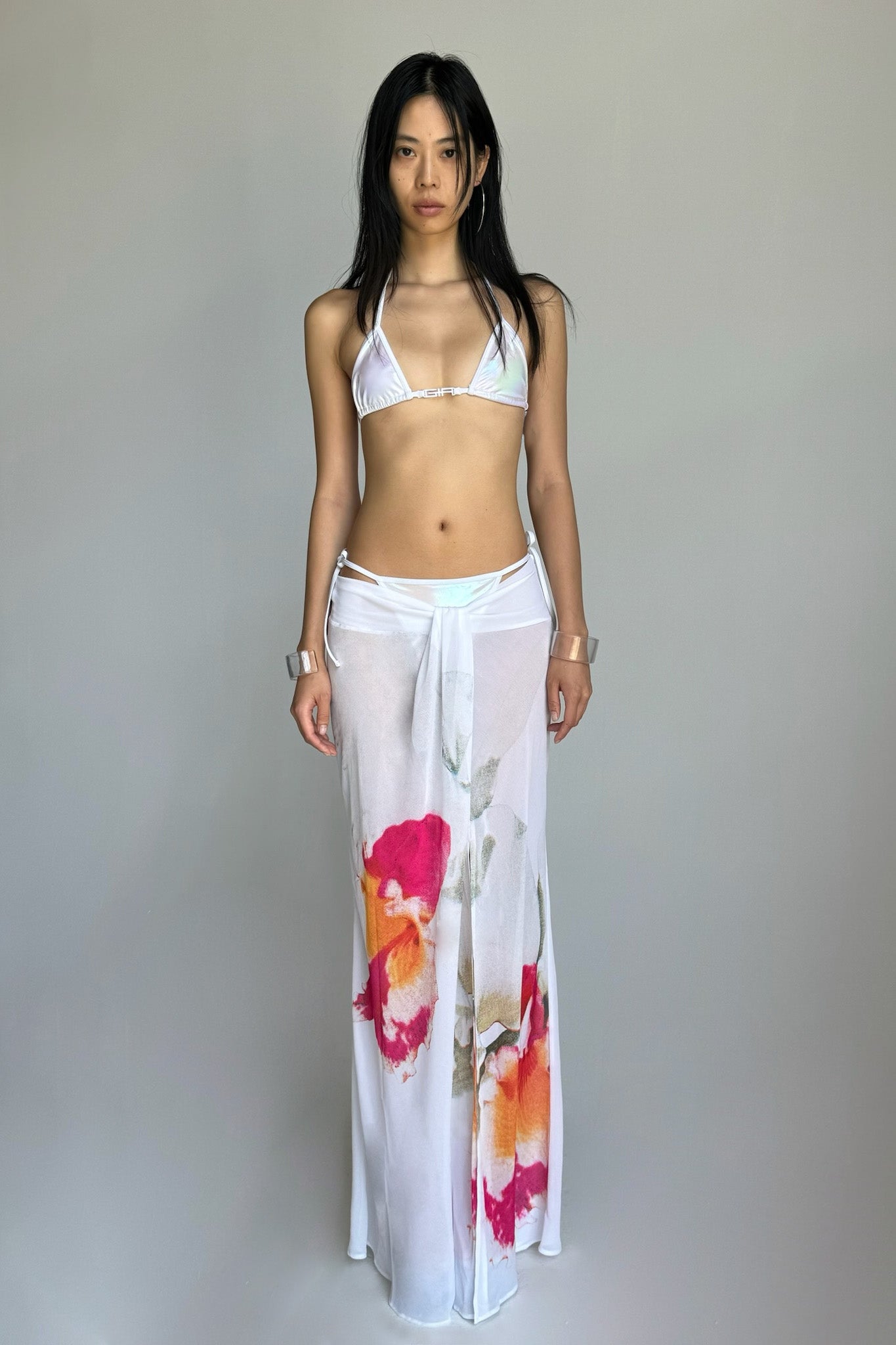 A woman stands against a plain background wearing an I.AM.GIA white bikini top and the PURA MAXI SKIRT - WHITE: FLORAL. She has long, straight black hair and a neutral expression, accessorizing with white wristbands.