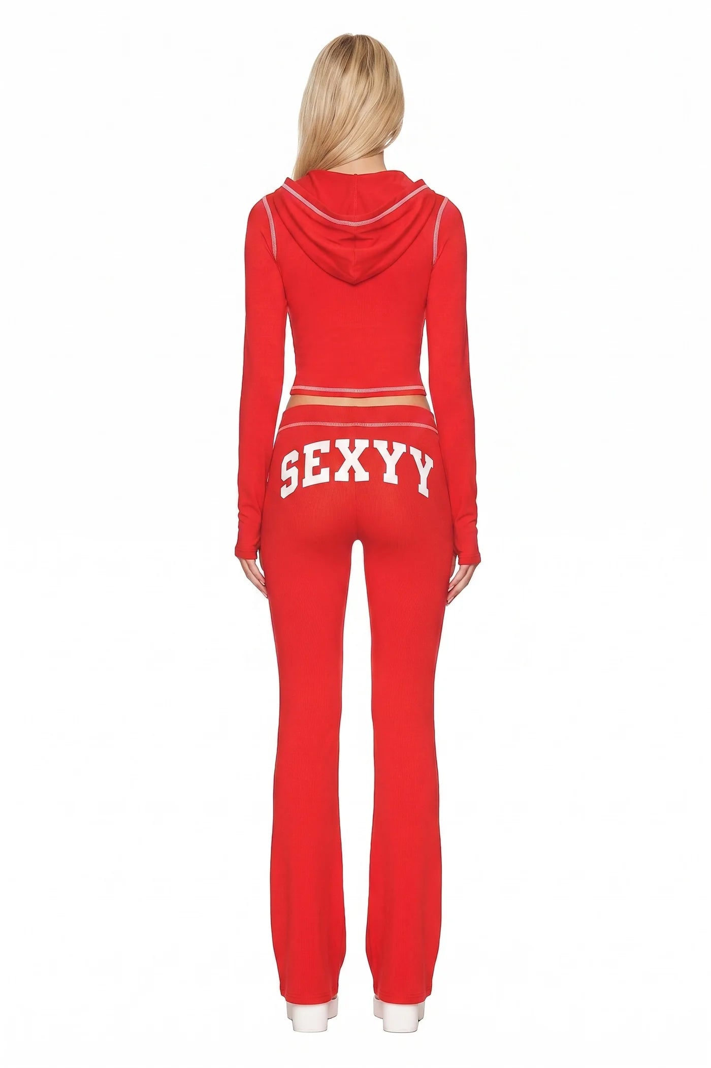 A person with long blonde hair, seen from behind, wears the I.AM.GIA X SEXYY RED BLARE HOODIE in red, featuring bold white "SEXY" lettering across the back of the pants. The vibrant outfit pops against a plain white background.