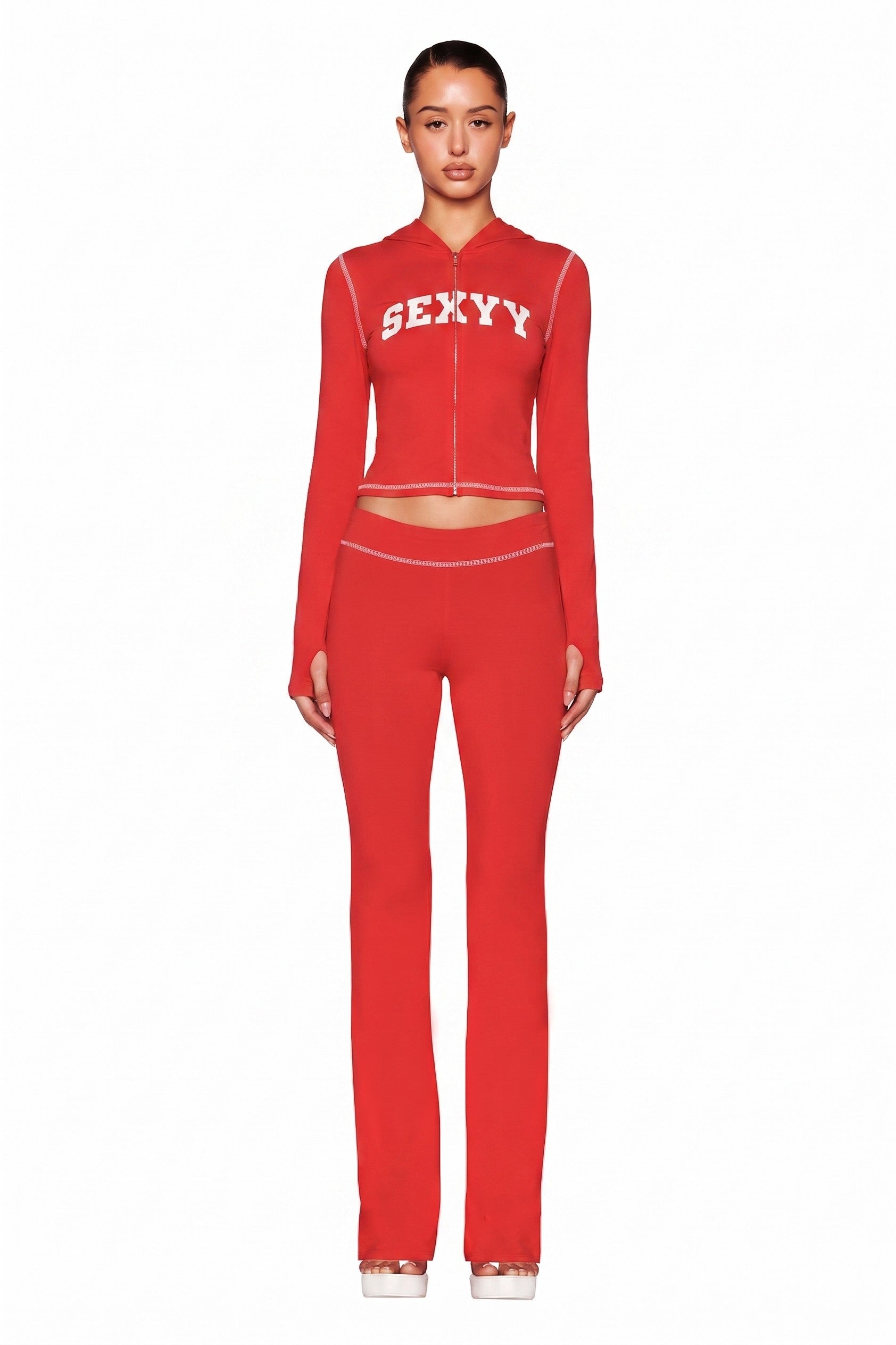 A woman faces forward in a fitted red I.AM.GIA X SEXYY RED BLARE HOODIE with "SEXYY" in white and matching high-waisted red pants with white stitching, standing against a plain white background.