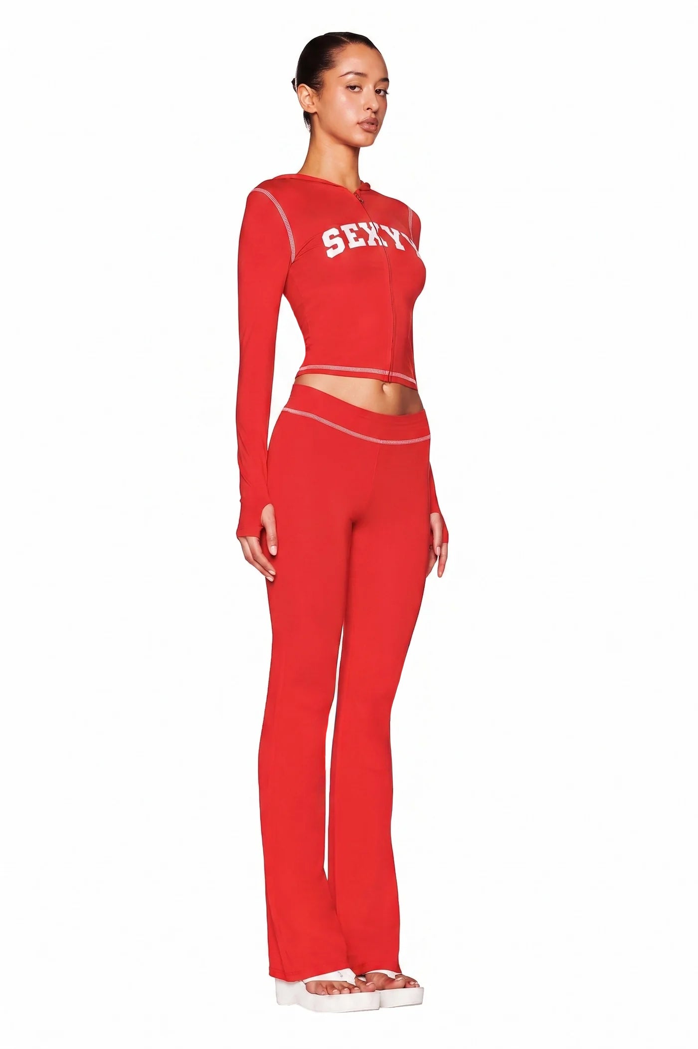 A woman stands in profile against a plain white background wearing the I.AM.GIA X SEXYY RED BLARE HOODIE in red, styled as a fitted two-piece with flared pants, white platform sandals, and "SEXY" on her top.