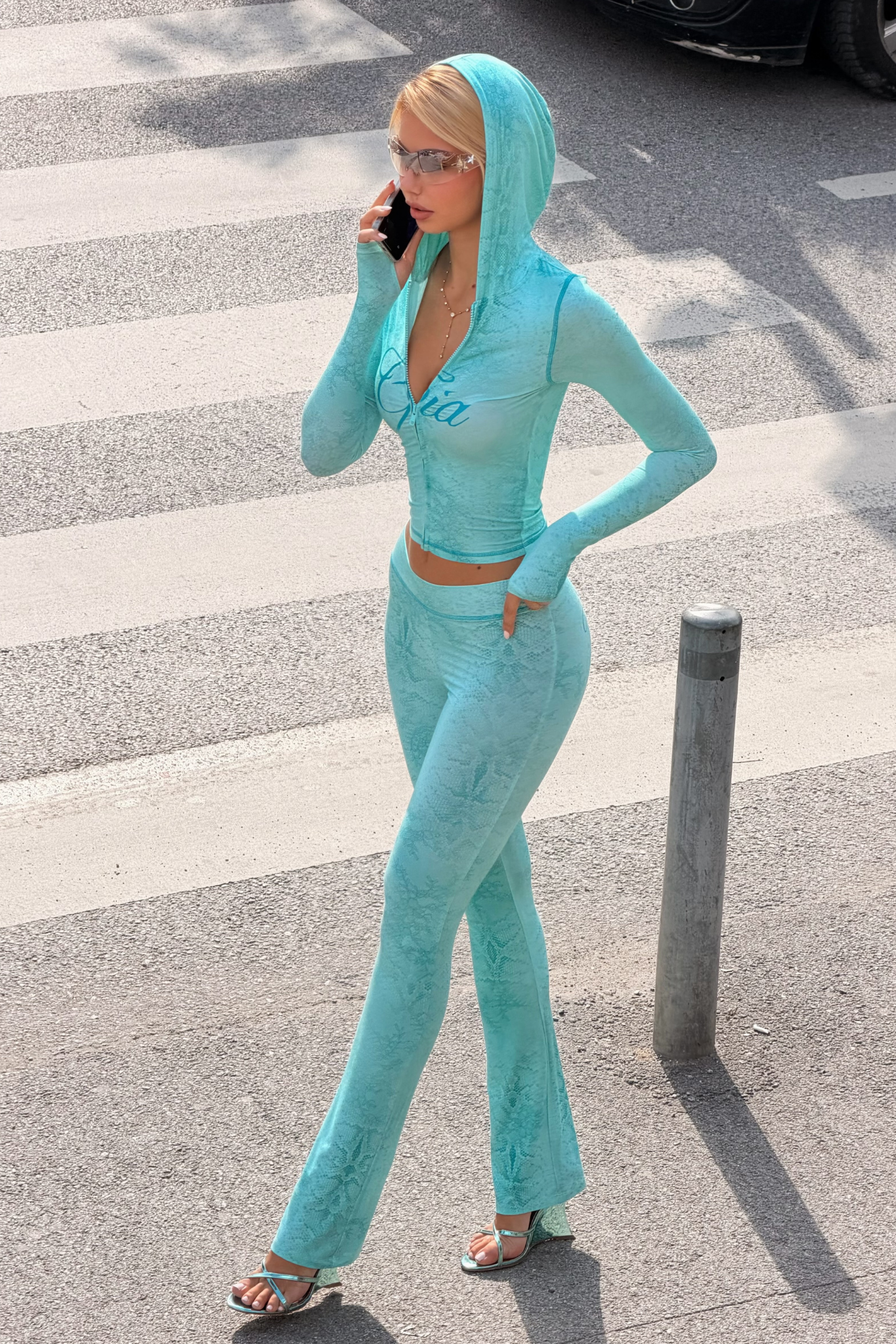 Wearing I.AM.GIA’s BLARE TRACKPANT in Teal Serpentine and a light blue hooded outfit, a person crosses the street near a metal post on a sunny day. They wear sunglasses and talk on the phone as they walk outdoors.