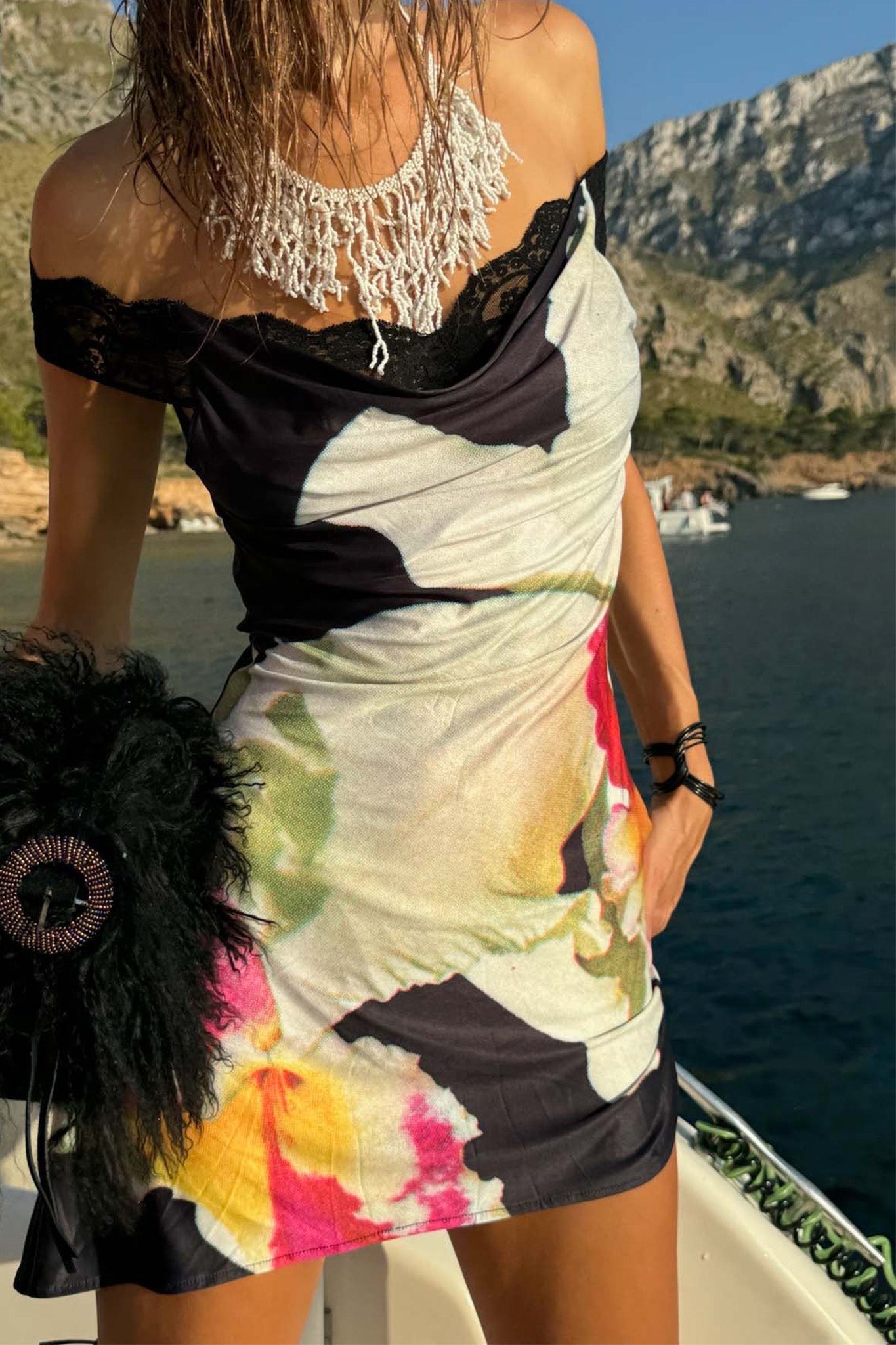A woman stands on a boat with rocky hills and water behind her, wearing the I.AM.GIA THEODORA MINI DRESS in black, a statement necklace, and holding a black feathered bag.