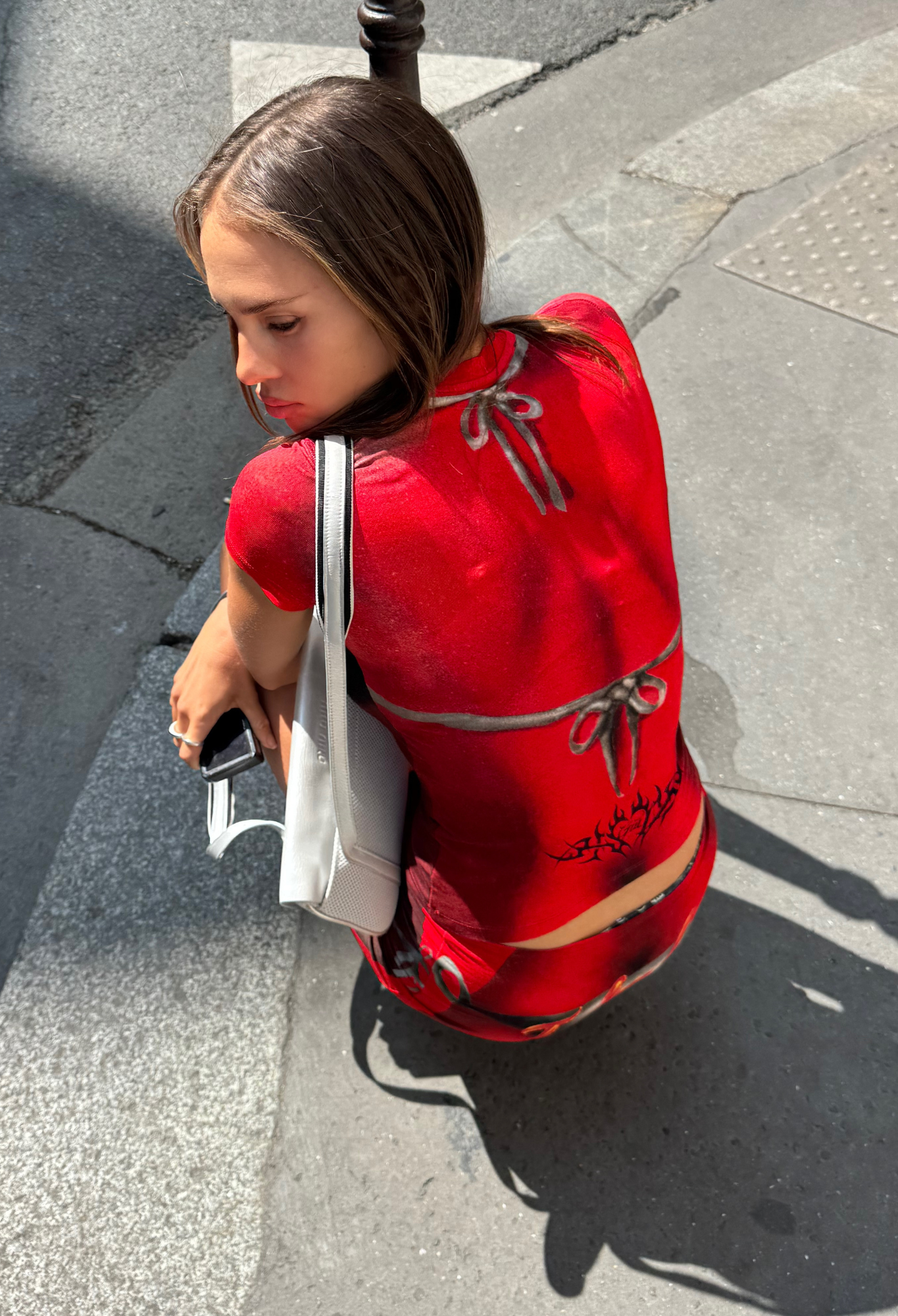 Wearing the I.AM.GIA T-REX SPRAY BIKINI BLARE TEE in red, a person crouches on a city sidewalk with a white bag and phone in hand, seen from above.