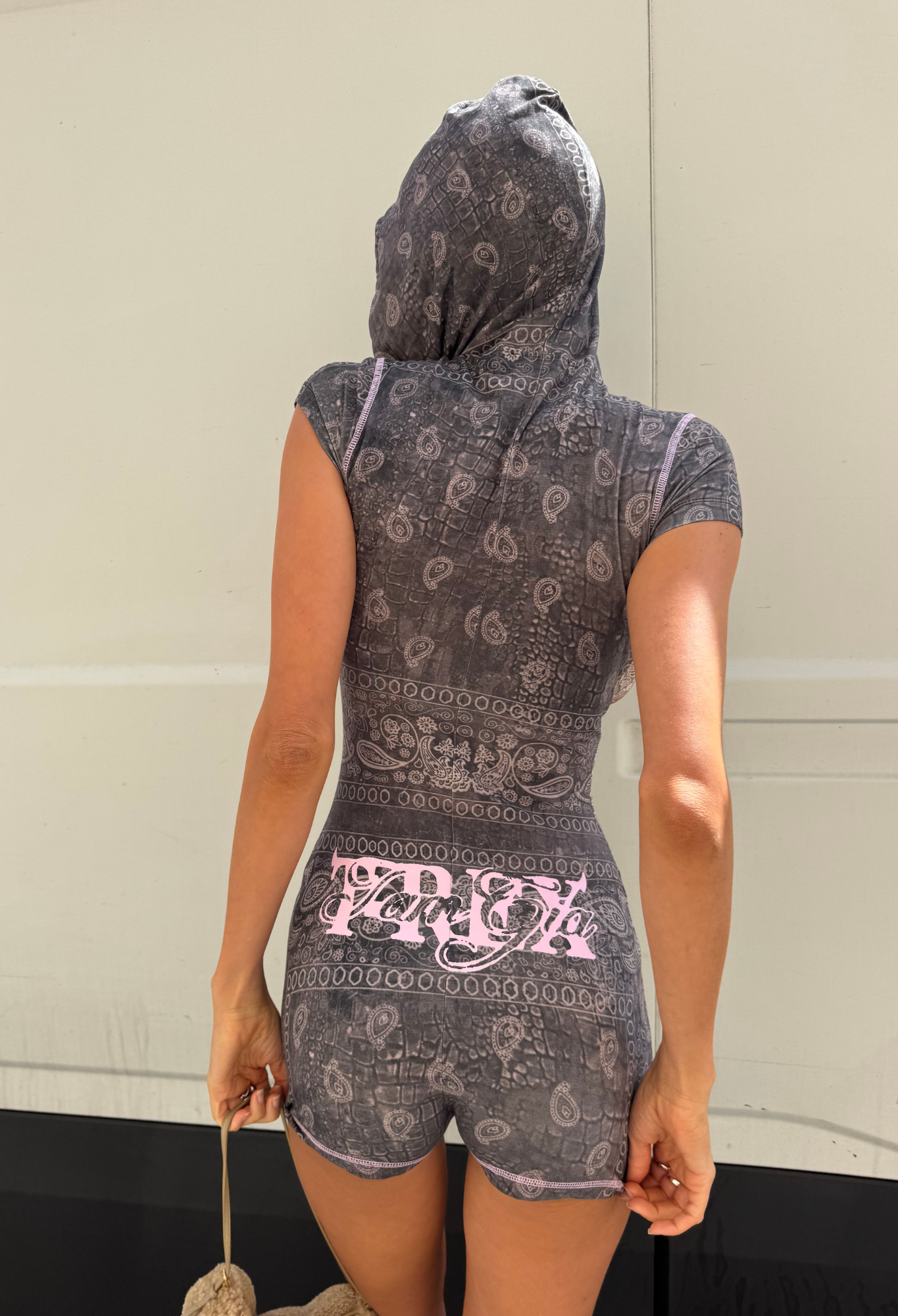 A person stands facing away, wearing the I.AM.GIA T-REX BLARE SHORT SLEEVE ROMPER in Charcoal Bandana, featuring a hood and pink branded accents, while holding a fuzzy accessory by their side against a plain light-colored background.